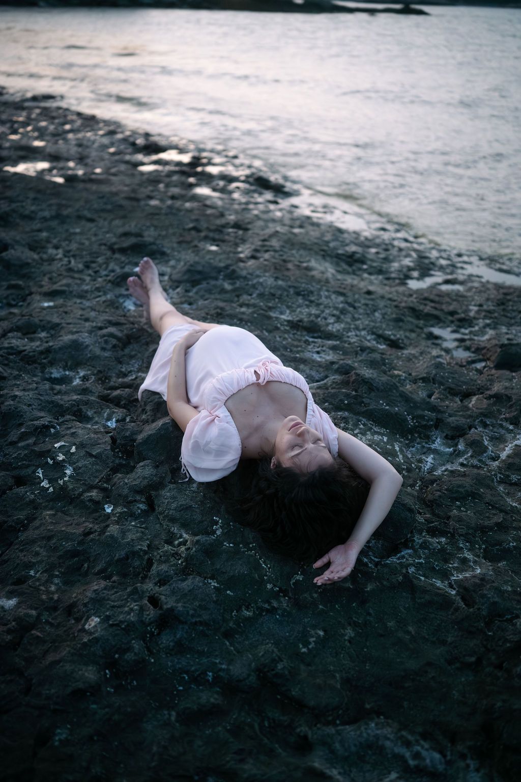 A figure in white lies on a dark rocky beach at dusk creating a dramatic contrast with the moody surroundings.