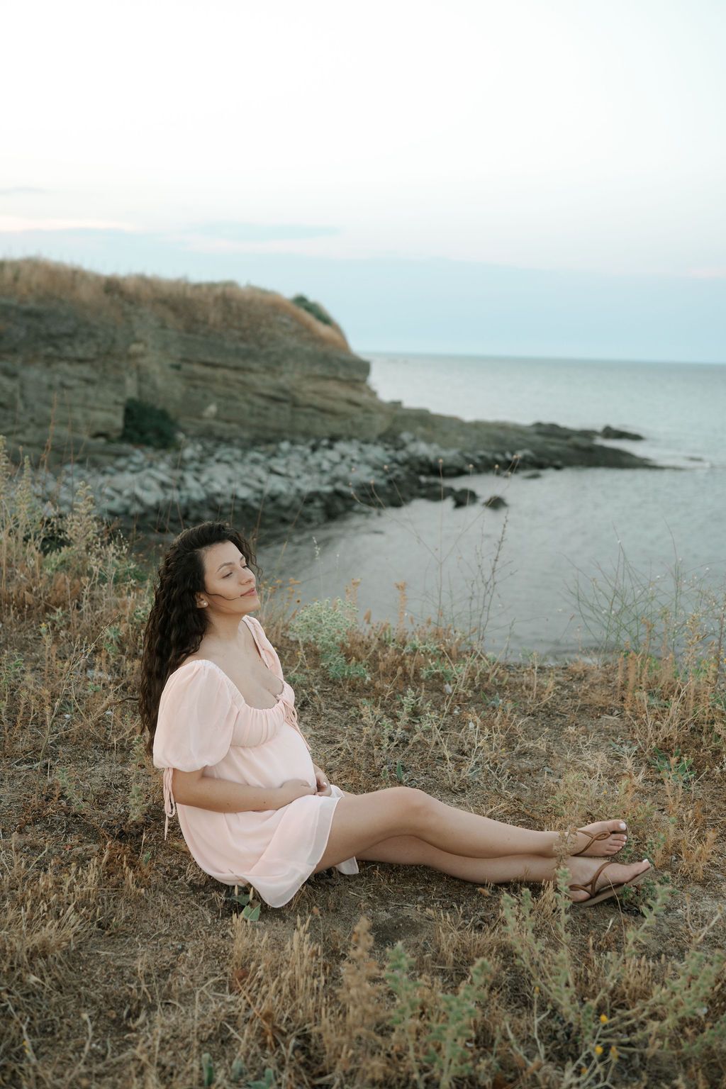 A person in a pink dress reclines on a coastal cliff overlooking the ocean during golden hour.