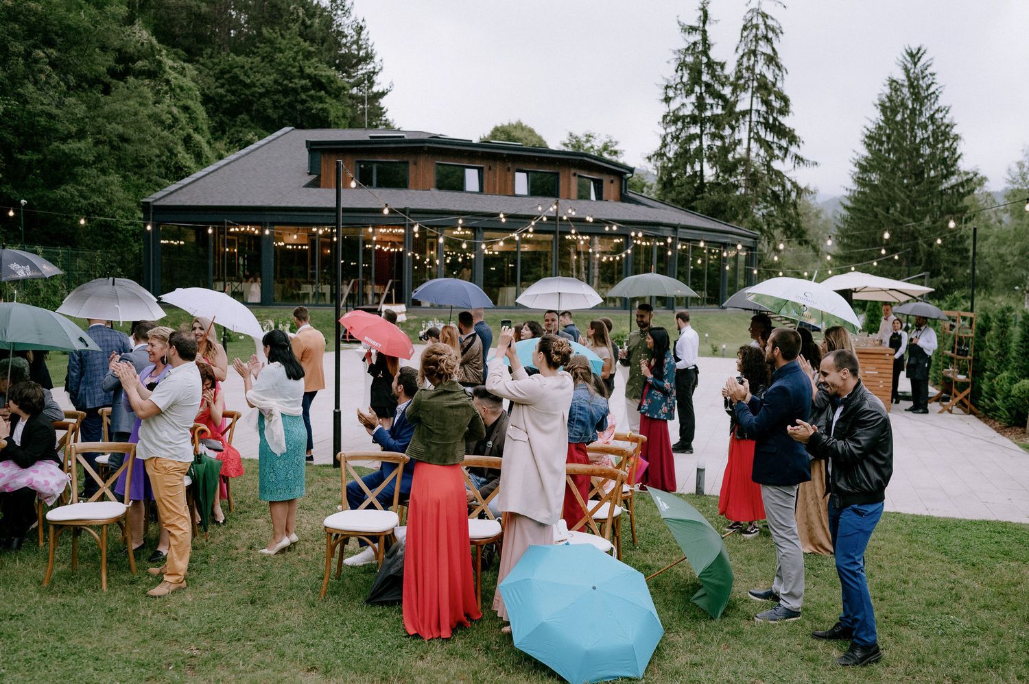 Panoramic view of outdoor wedding guests mingling under evergreen trees during a rainy reception celebration.
