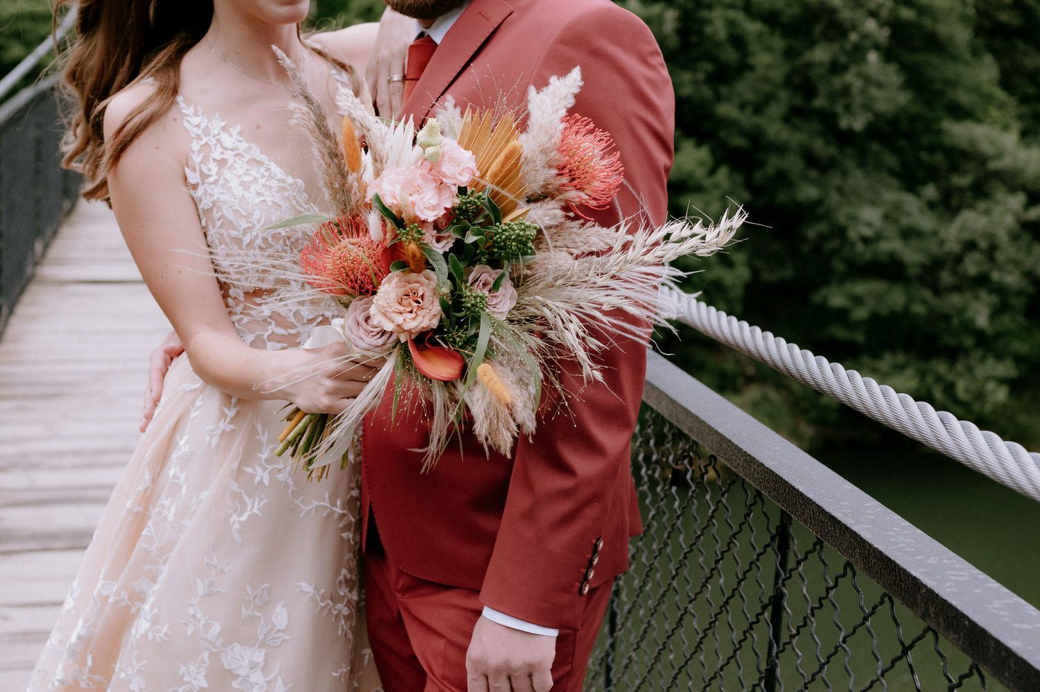 A wedding couple on a bridge holding a romantic bouquet with coral roses, pampas grass and dried flowers.