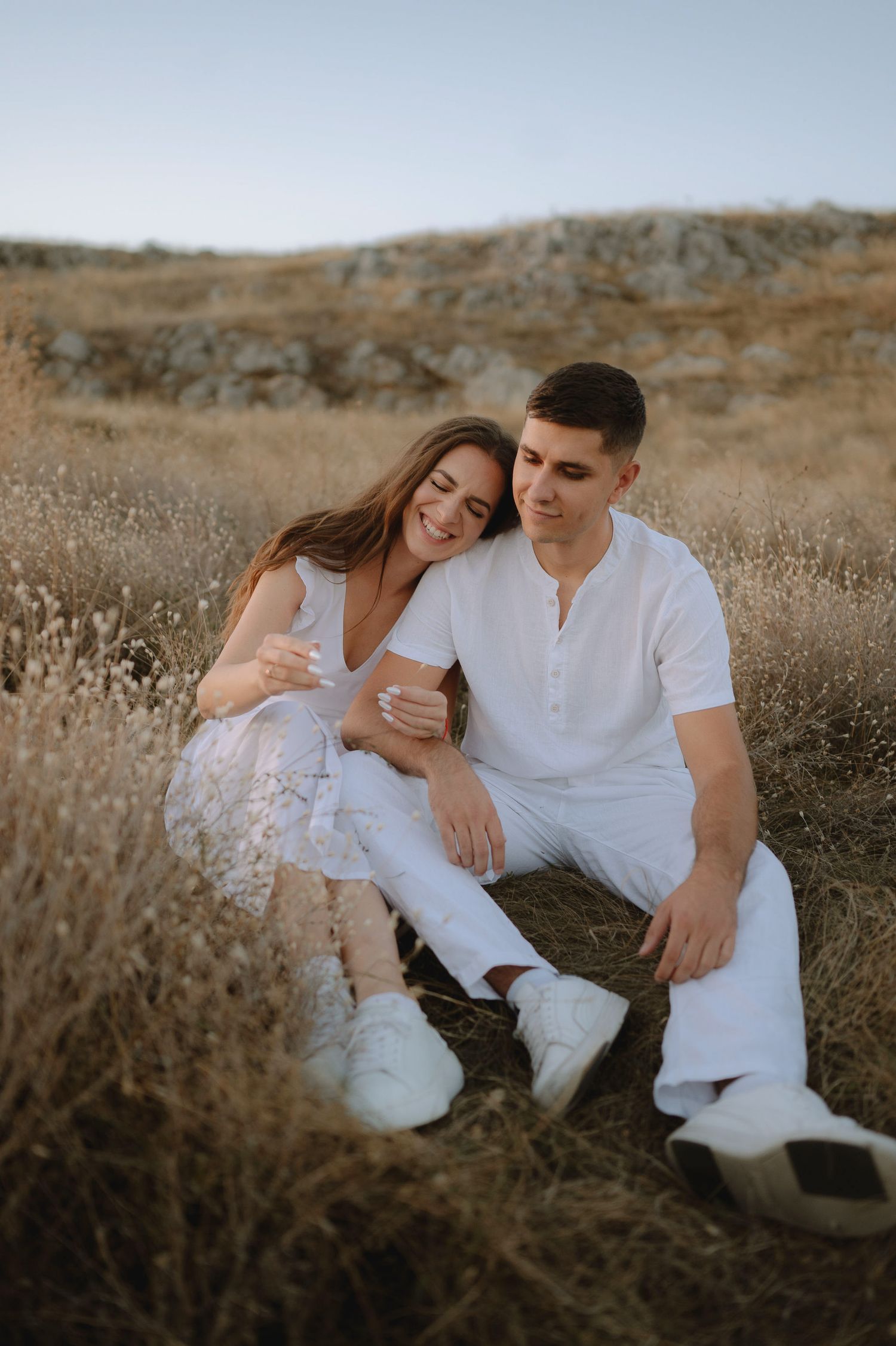 A couple in white clothing sits together intimately in a golden field during sunset.