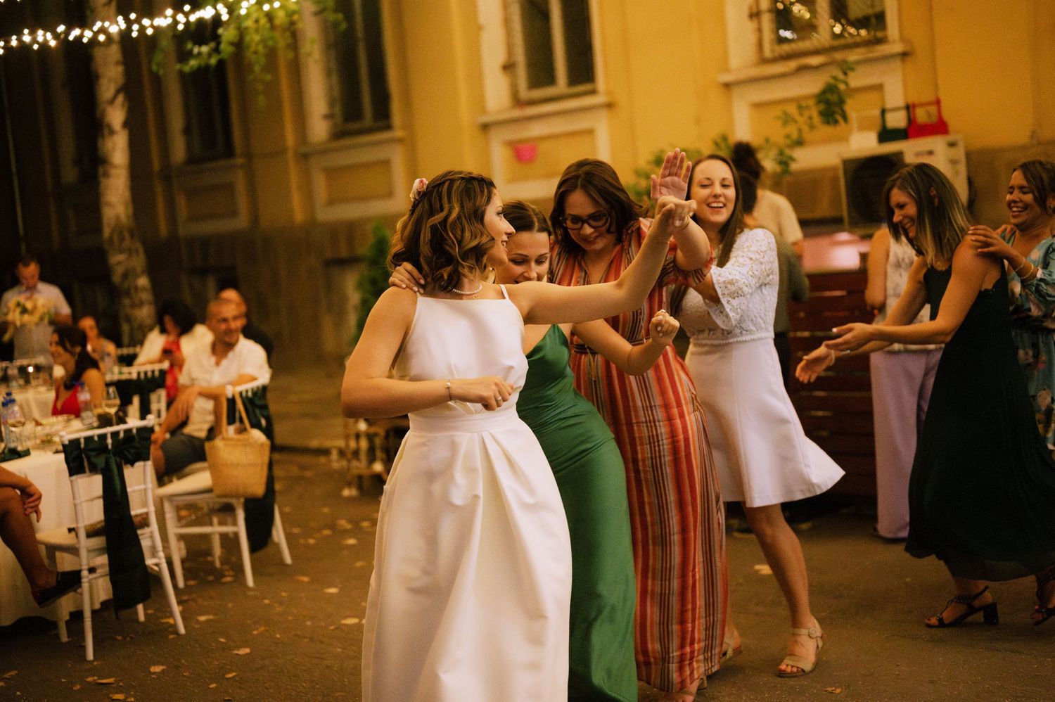 Wedding guests dance together at an outdoor reception venue illuminated by string lights and warm ambient lighting.