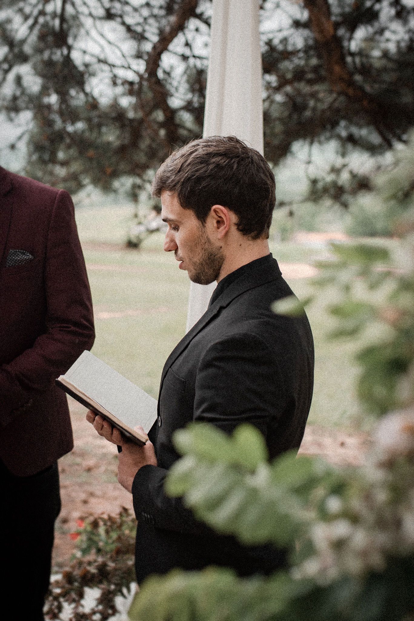 A person in a black suit reads from papers during an outdoor ceremony framed by greenery and trees.