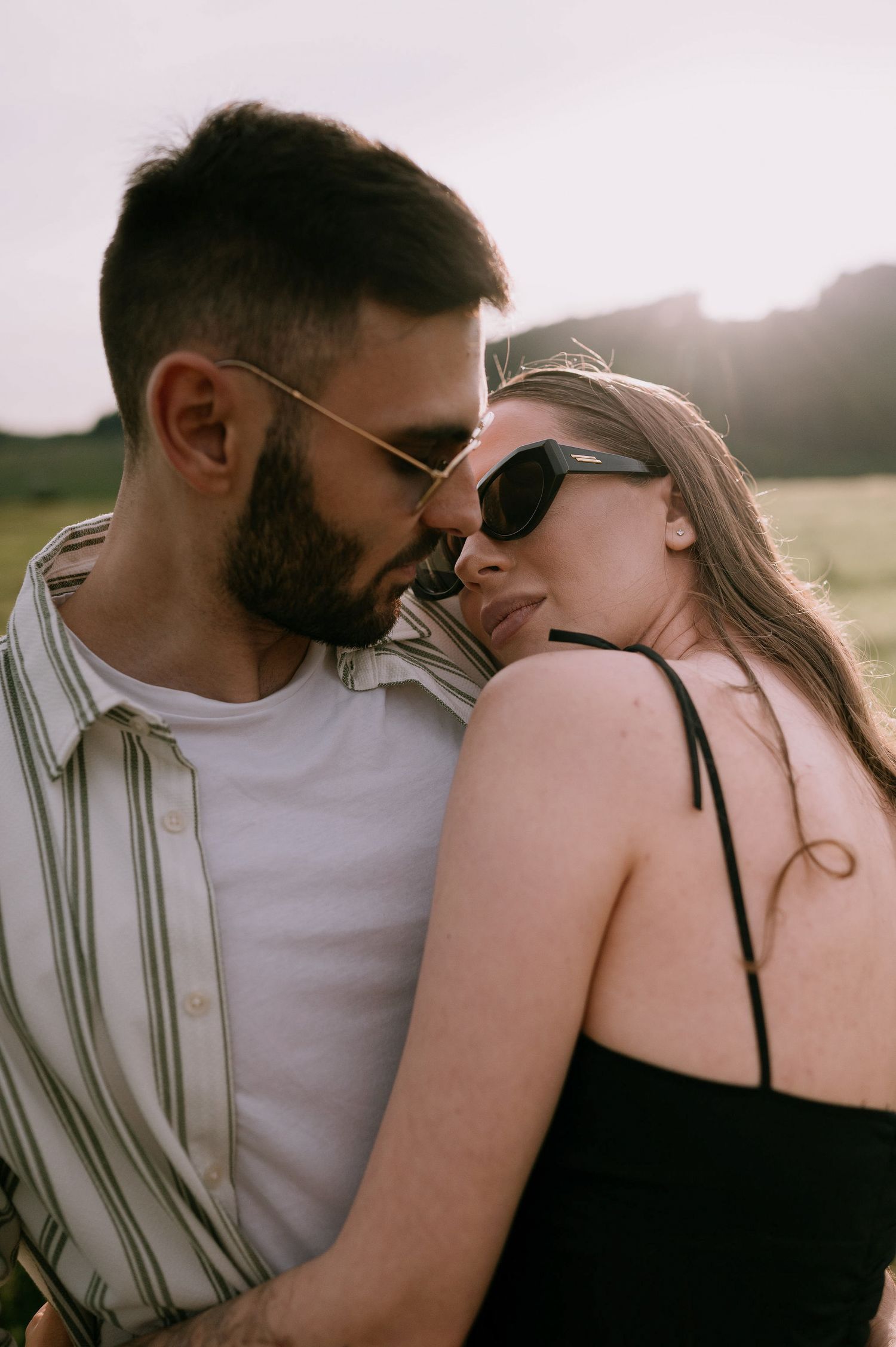 A tender moment between a couple in sunlit field wearing casual attire and sunglasses.