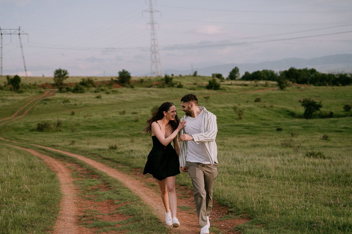 A couple shares a romantic moment together while walking along a dirt path through rolling green prairie fields.