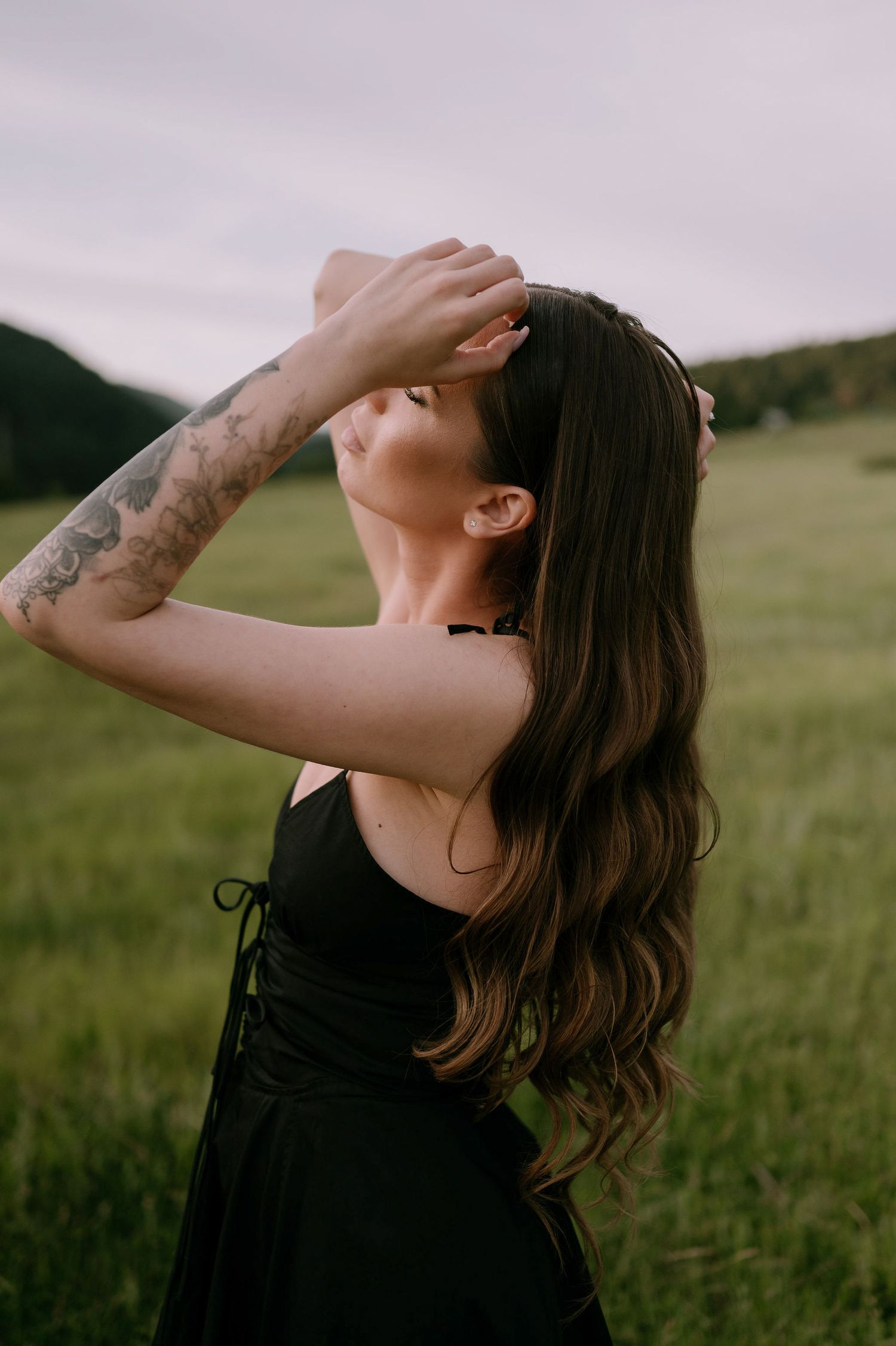 A person in a black tank top tilts their head back while standing in a grassy field at sunset.