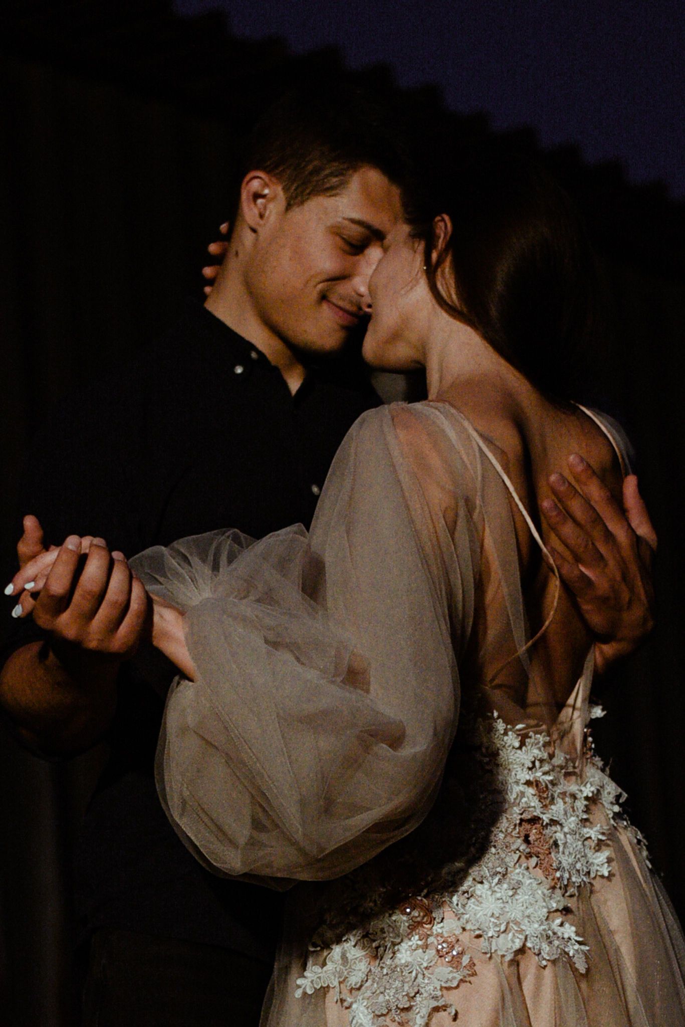 A romantic wedding couple embraces in a dark setting, with the bride wearing a delicate lace and tulle gown.