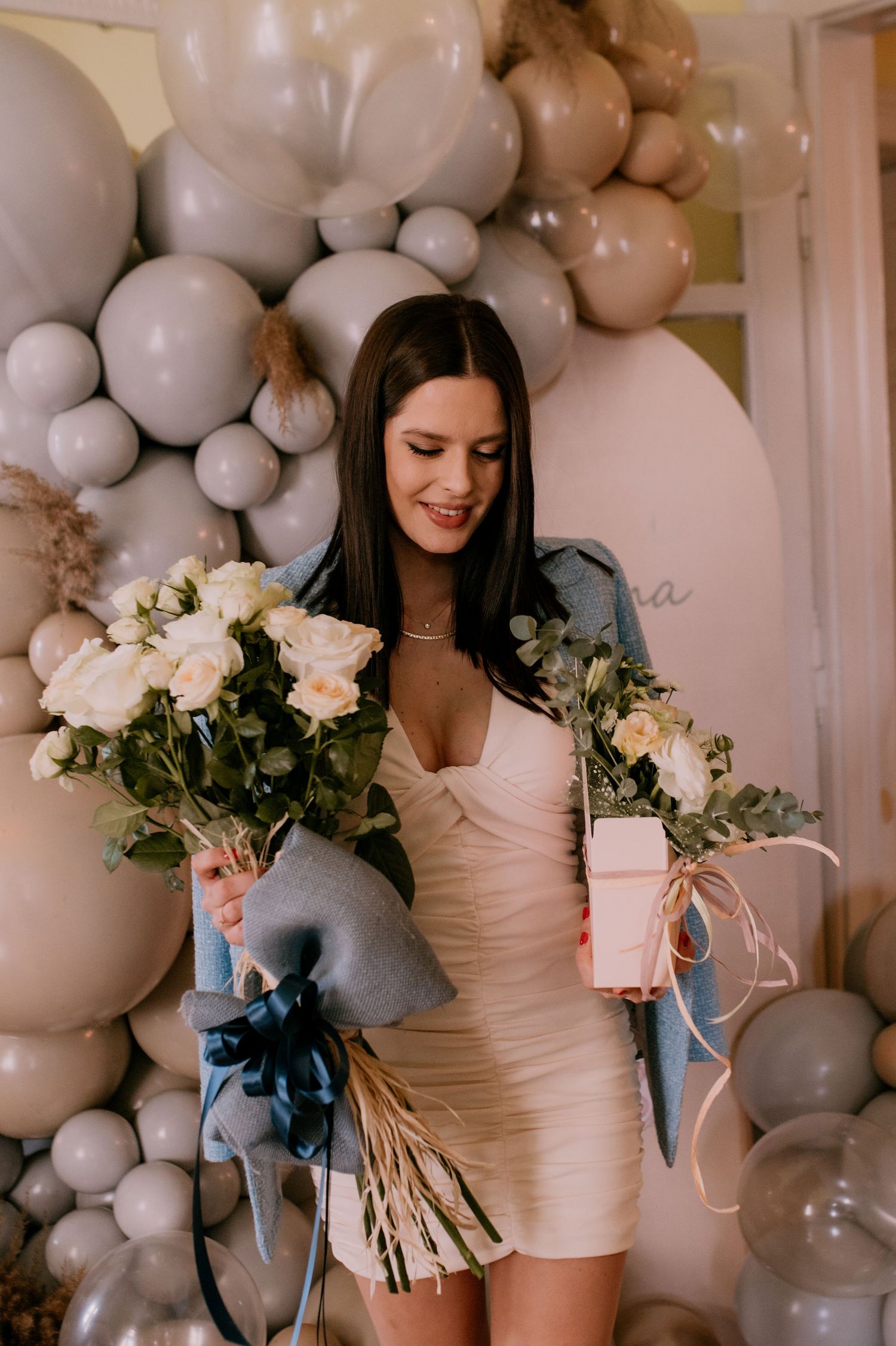 Woman in white dress holds white roses against a backdrop of elegant grey and white balloon decorations.