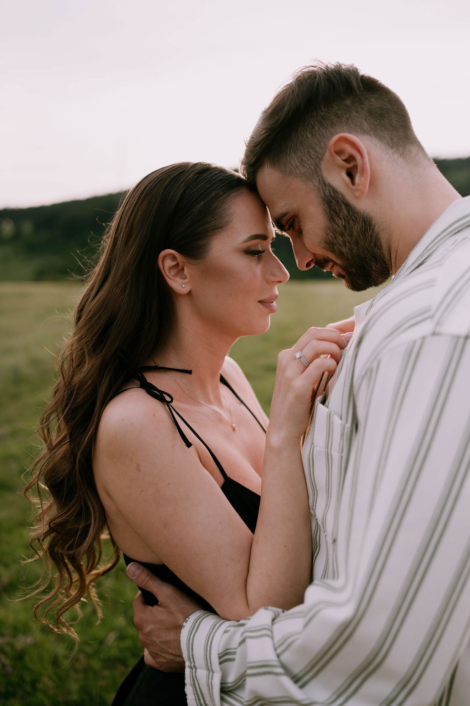 Intimate couple embraces in a field during sunset, with her in black and him in white stripes.