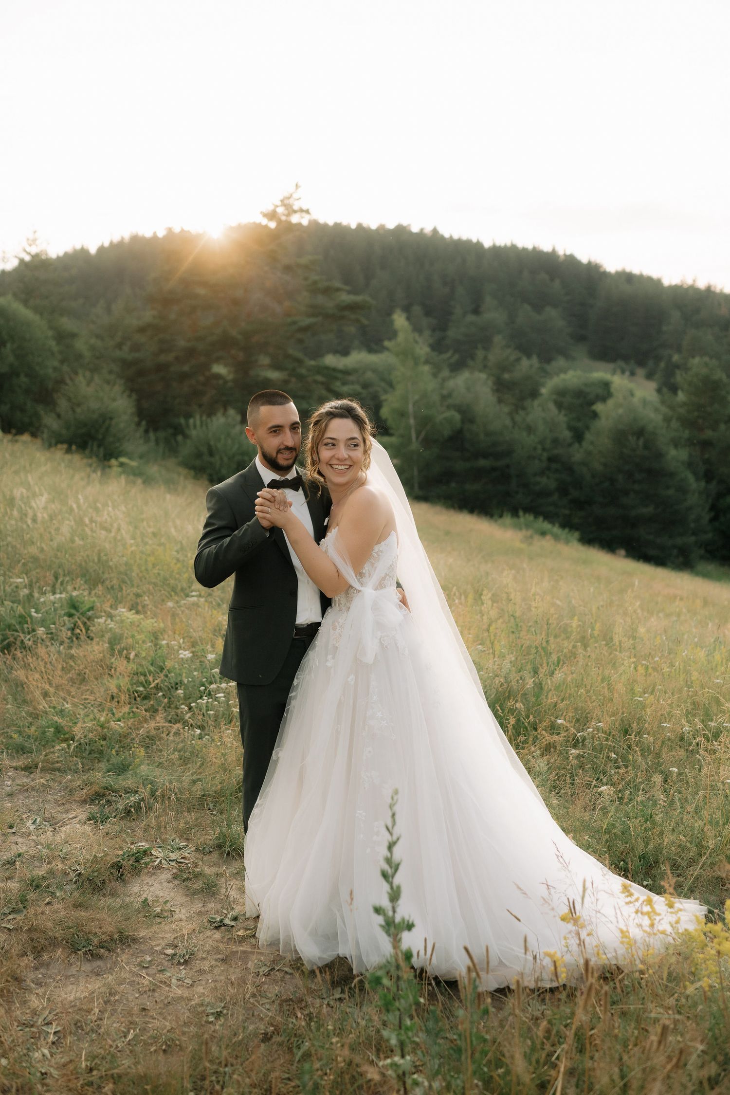A newlywed couple shares a romantic moment in a sunlit meadow at golden hour with mountains in the background.