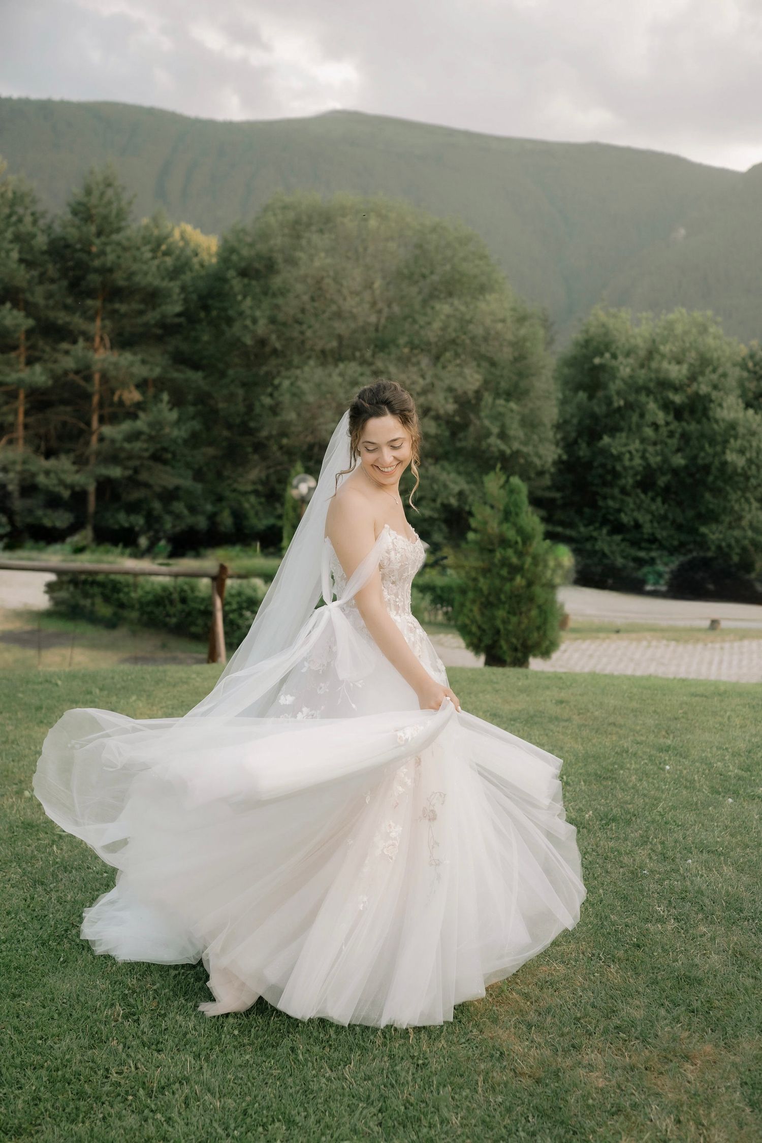 A bride in a flowing white wedding dress twirls gracefully on a grassy hillside with mountains in the background.