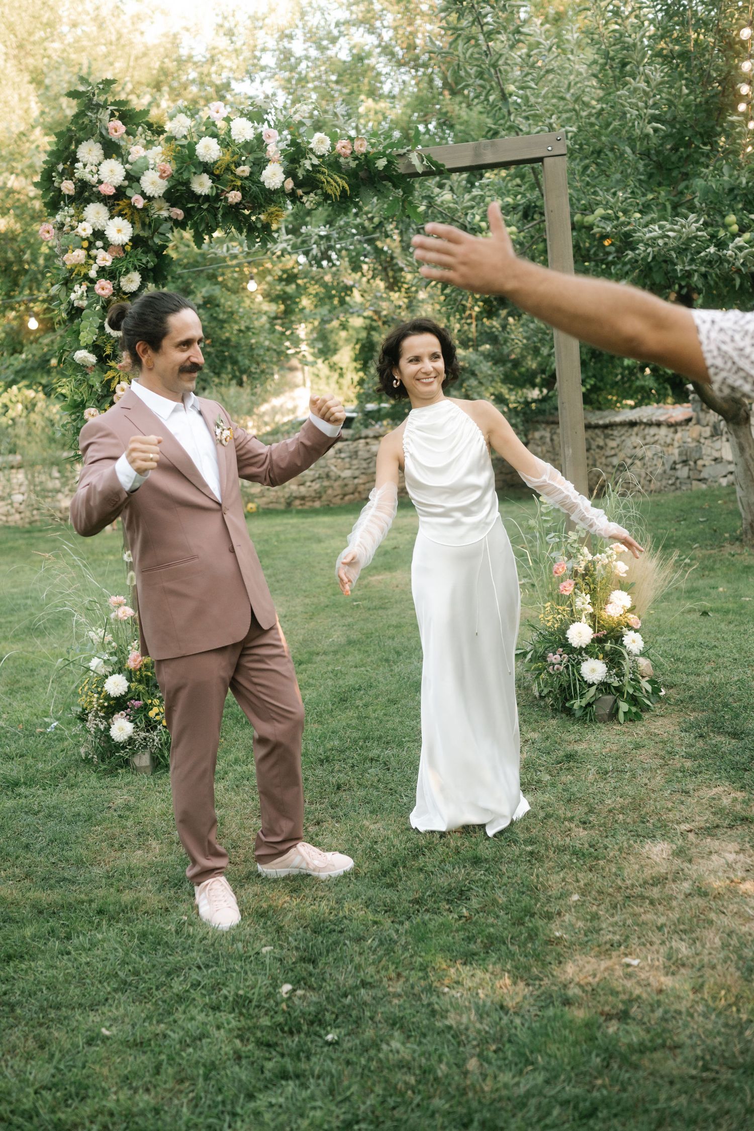 A couple in wedding attire dances playfully on grass with floral arrangements in the background.