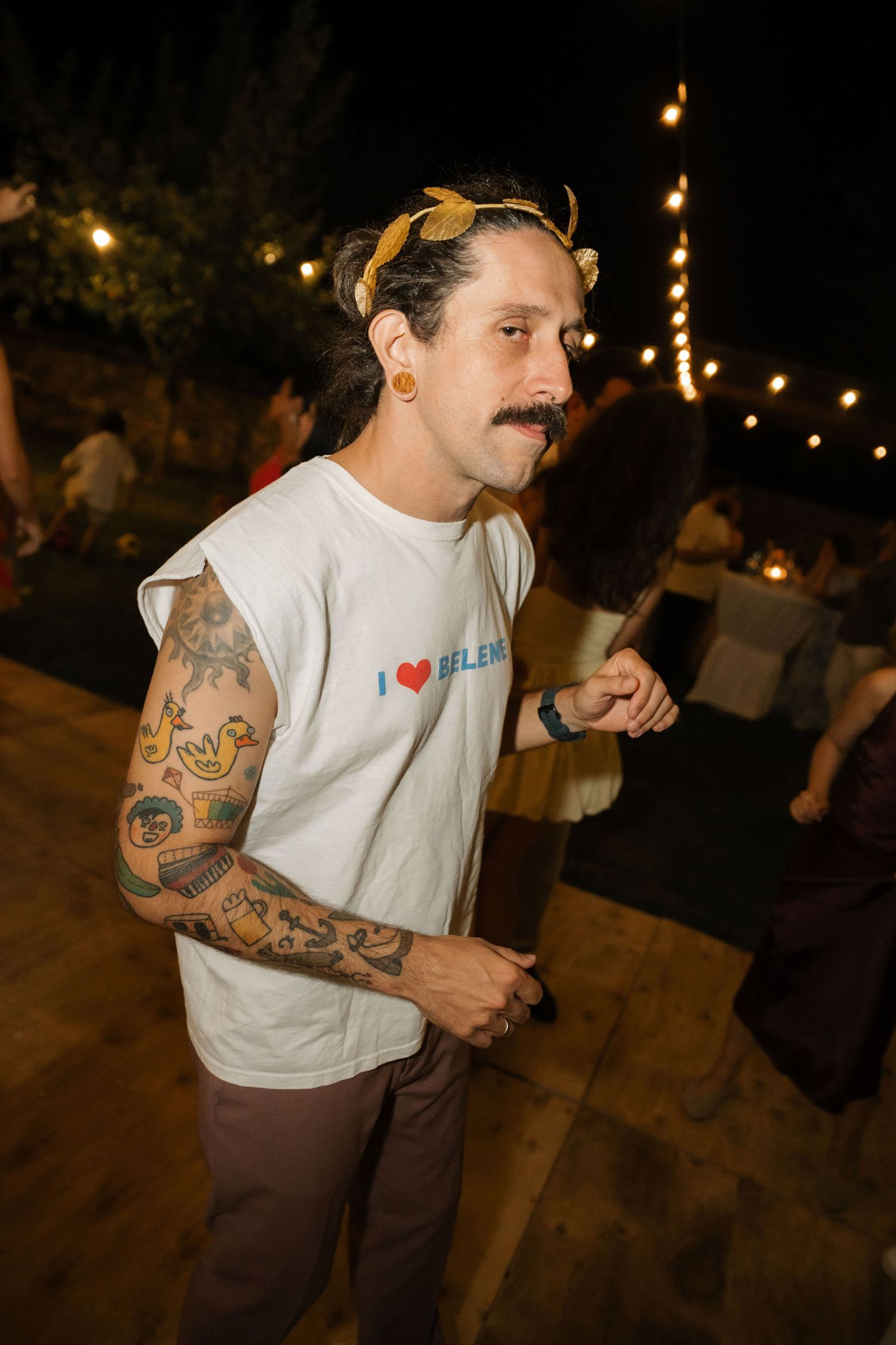 Person in white t-shirt with tattoos dancing at night under string lights at outdoor event.