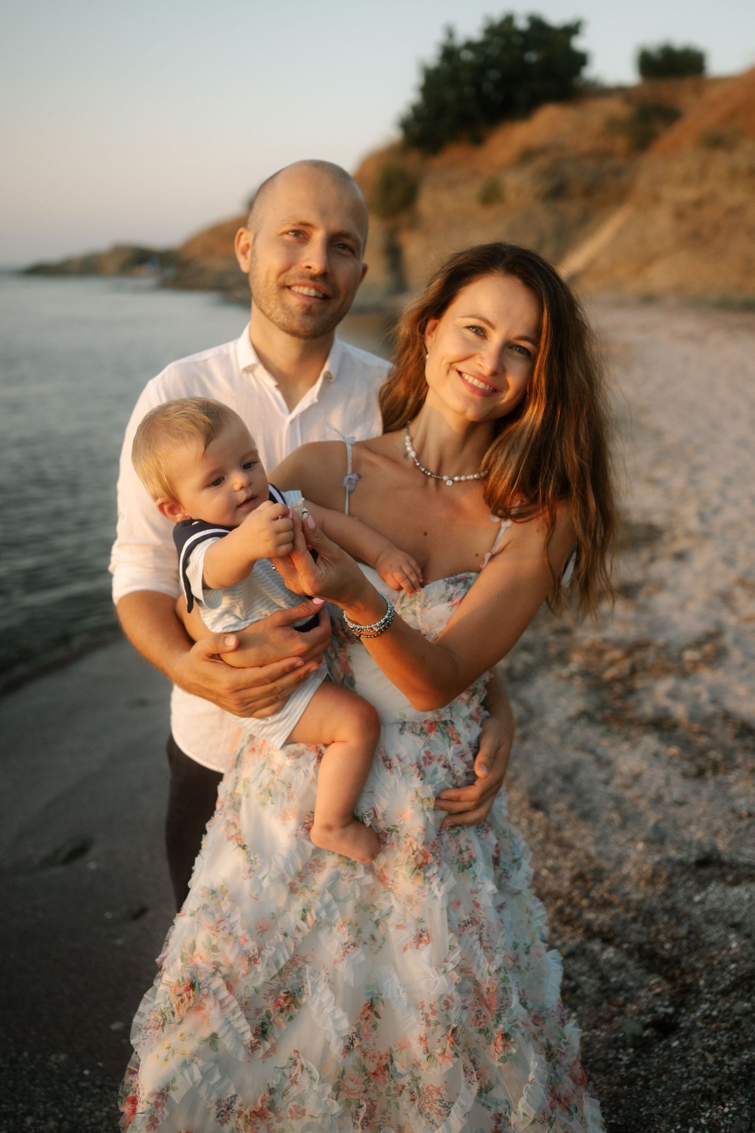 A family shares tender moments on a beach at sunset, with a floral dress and white shirt creating a romantic coastal scene.