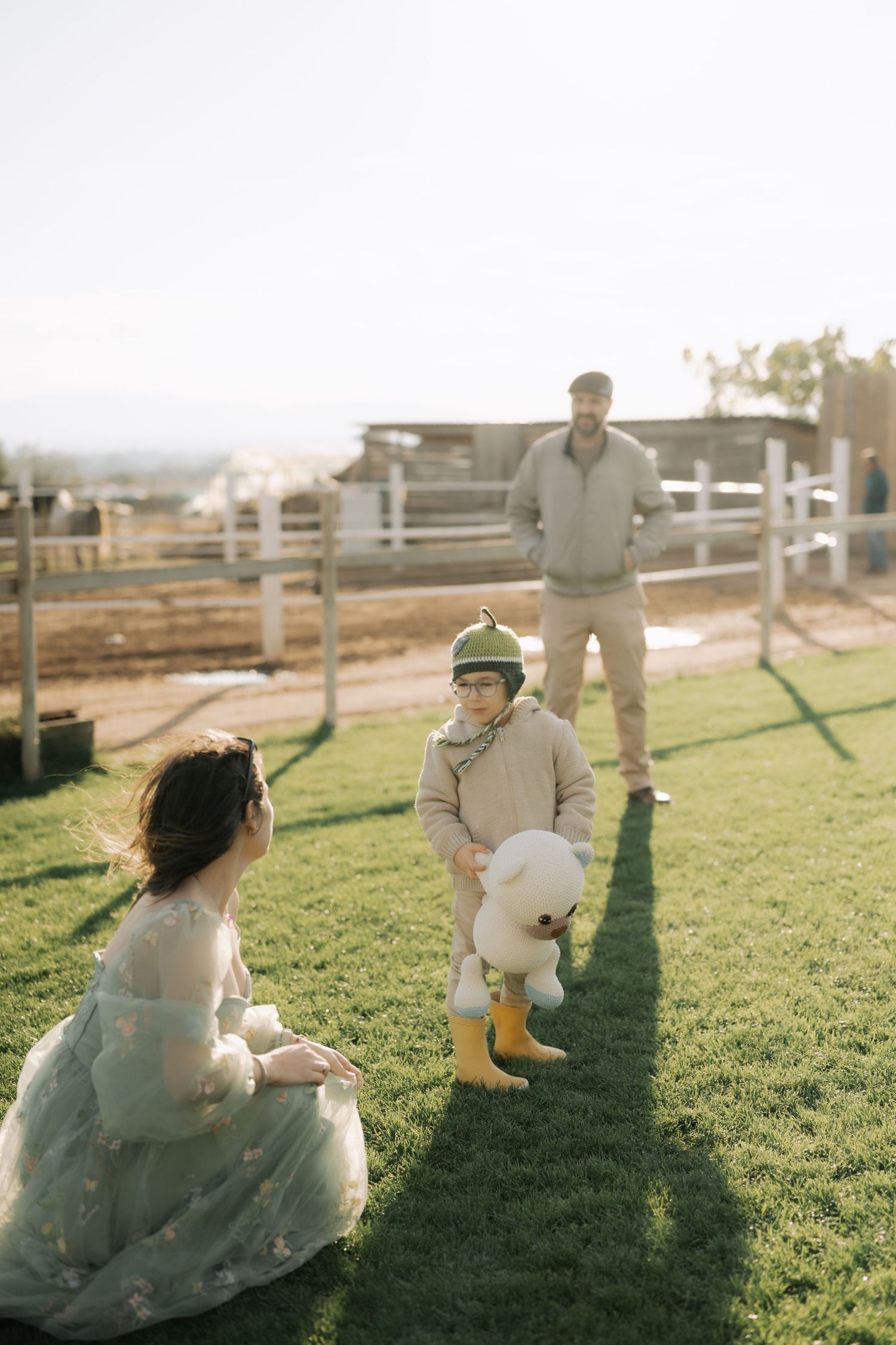 Family enjoying a sunny afternoon together on a grassy hill at a ranch with white fences in the background.