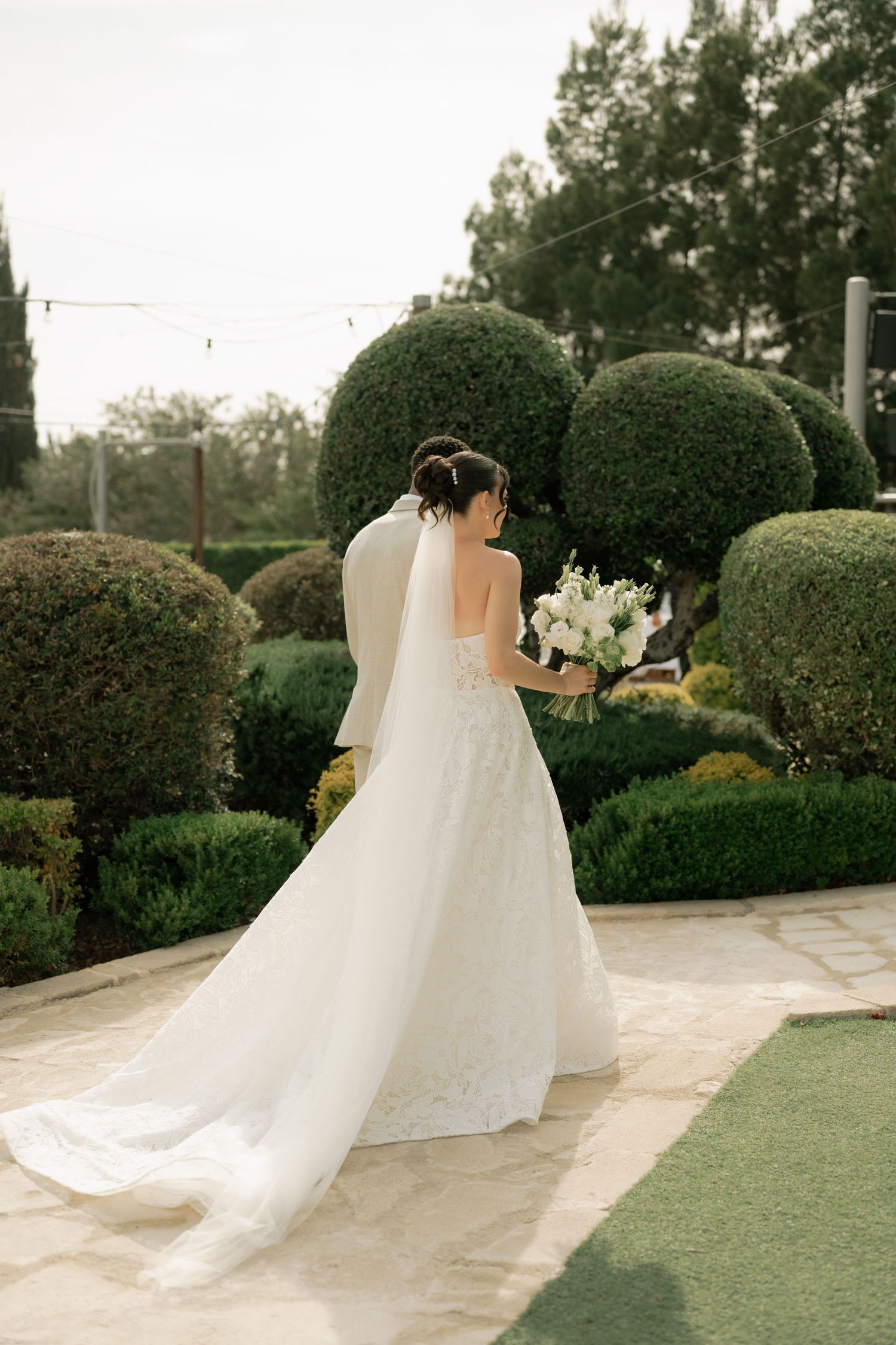 Bride in white lace gown and veil holds bouquet, walking through manicured garden with topiary bushes.