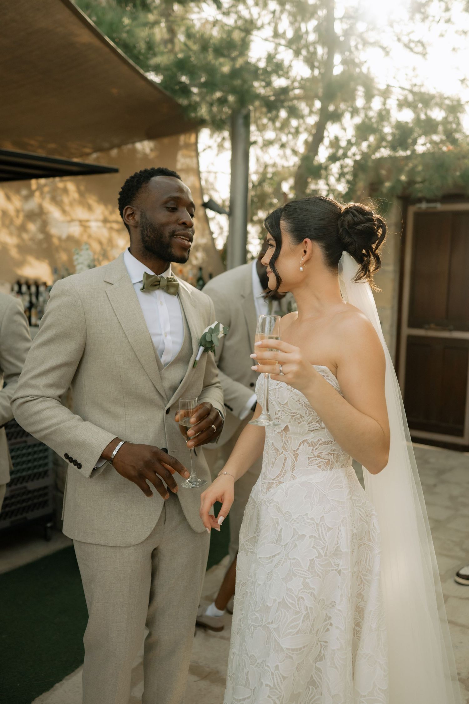 Bride and groom share a champagne toast outdoors, groom in beige suit with bow tie, bride in lace strapless gown with veil.