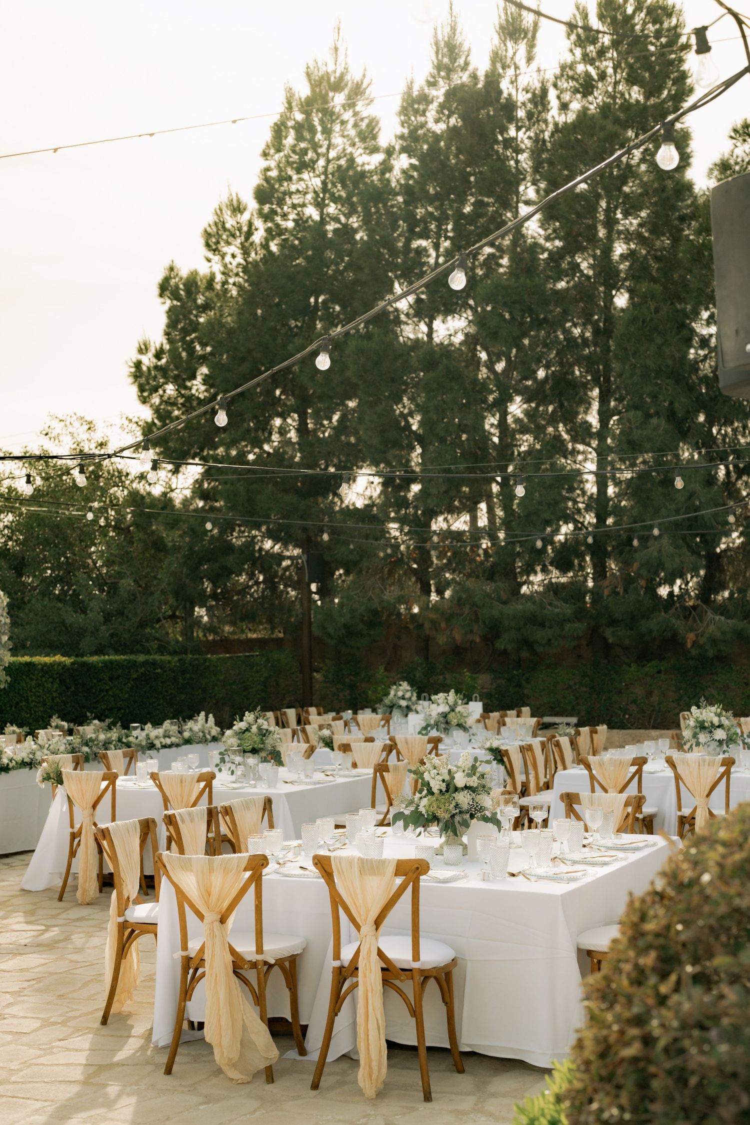 Elegant outdoor wedding reception with wooden cross-back chairs, white linen tables, floral centerpieces, and string lights among tall trees