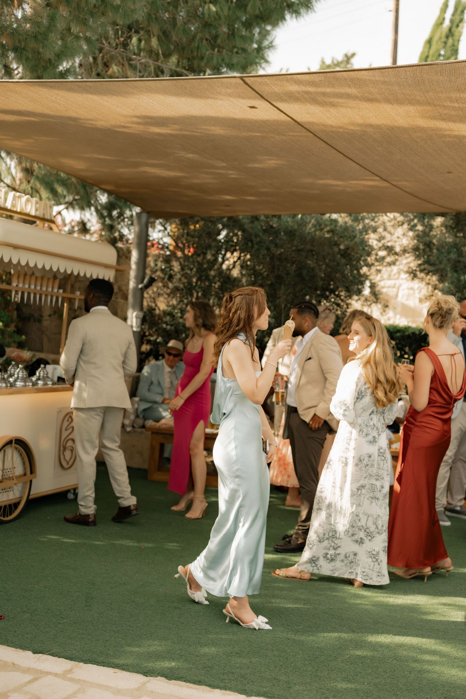 Guests mingle at an outdoor wedding reception under a tan canopy, wearing elegant attire including a blue satin dress.