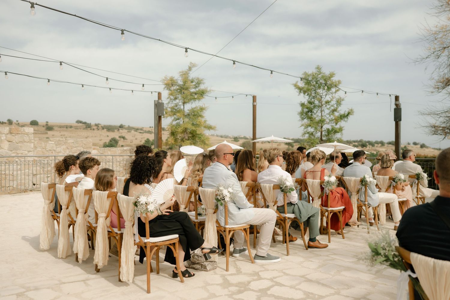 Guests seated in wooden chairs at an outdoor wedding ceremony on a stone patio with string lights overhead.