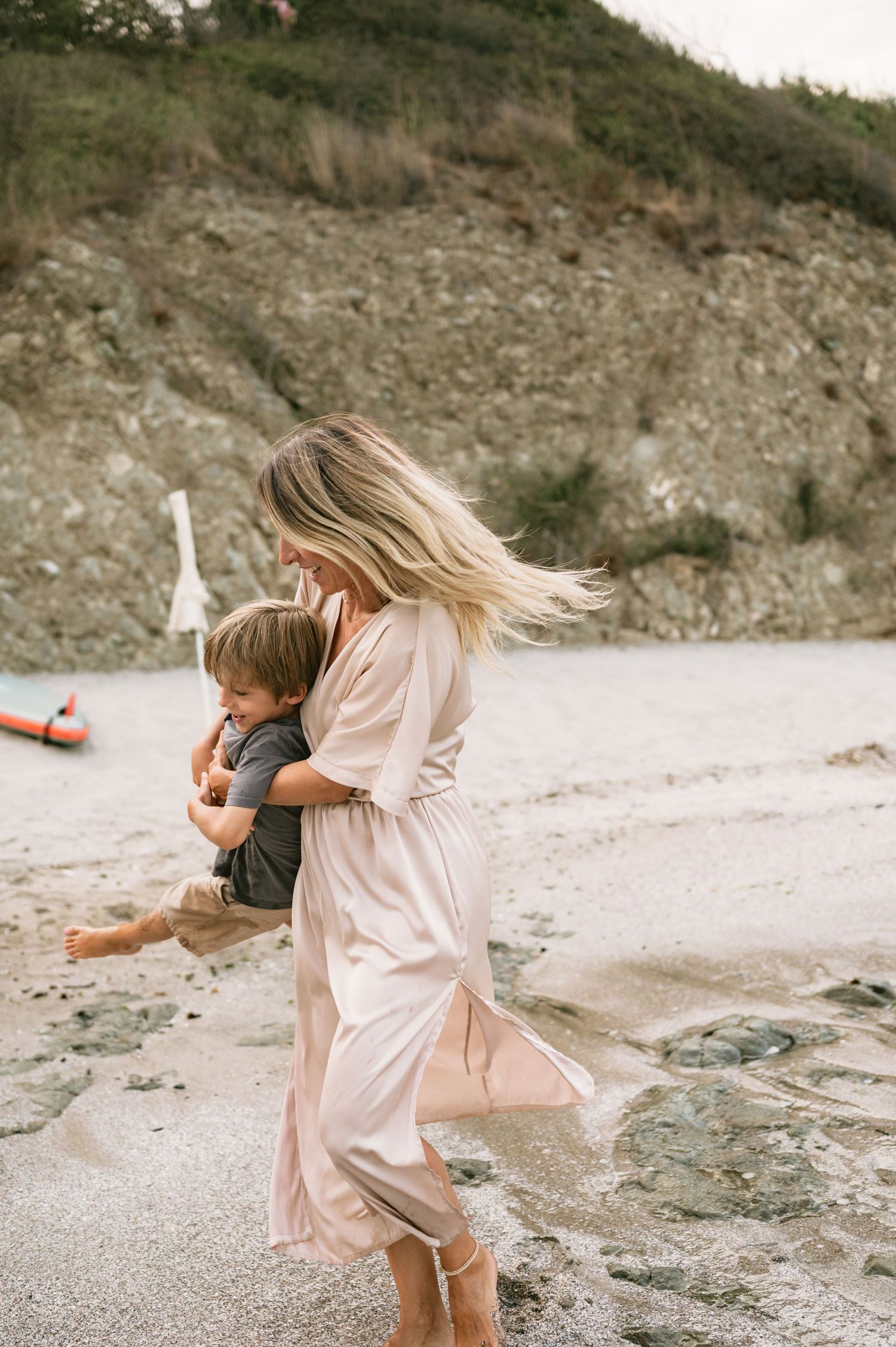 A woman in a flowing pink dress carries a young child on a sandy beach with rocky cliffs in the background.