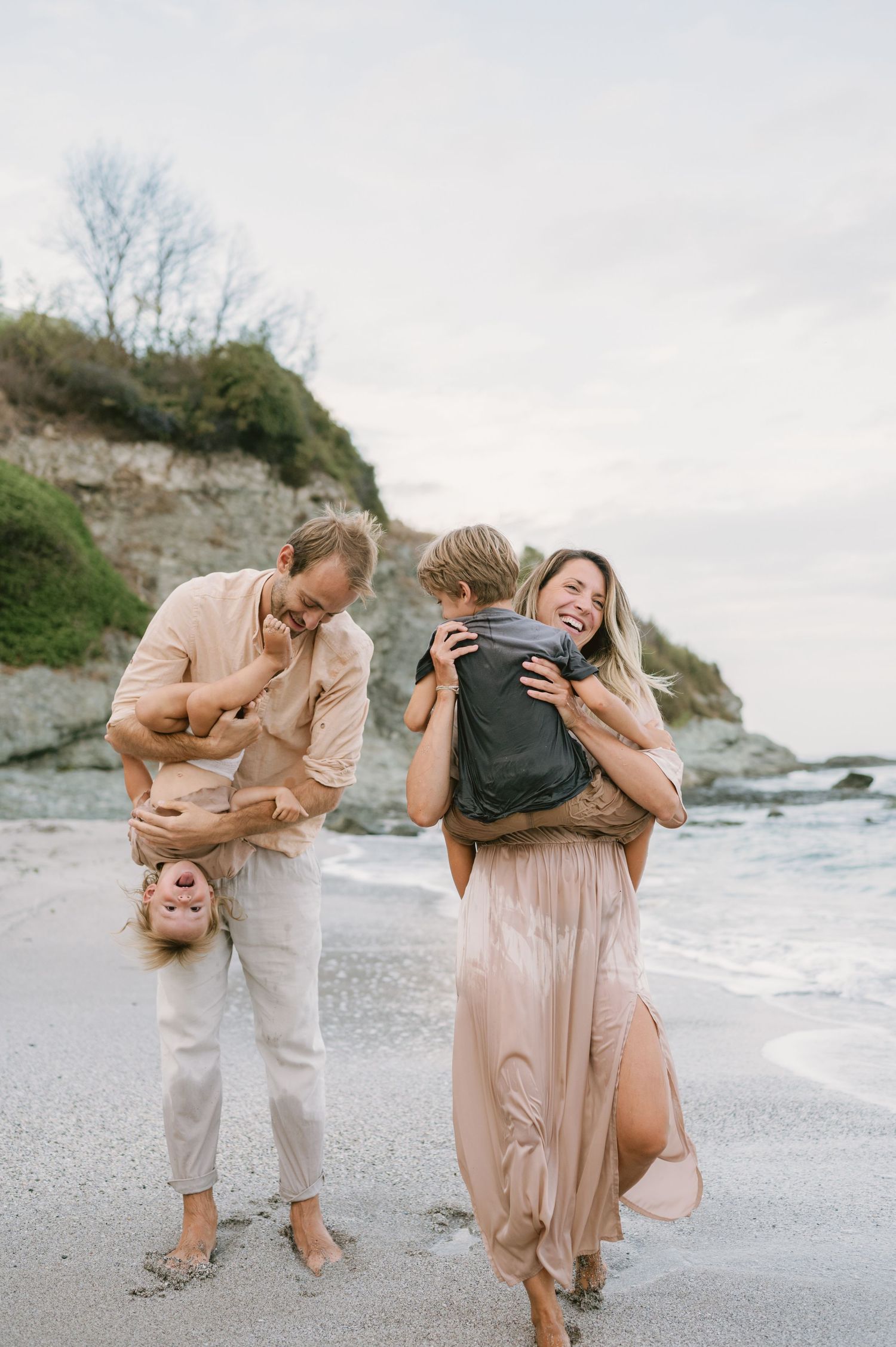 Family laughing on a beach, dad holding child upside down while mom hugs another child, sandy shore with cliffs behind.