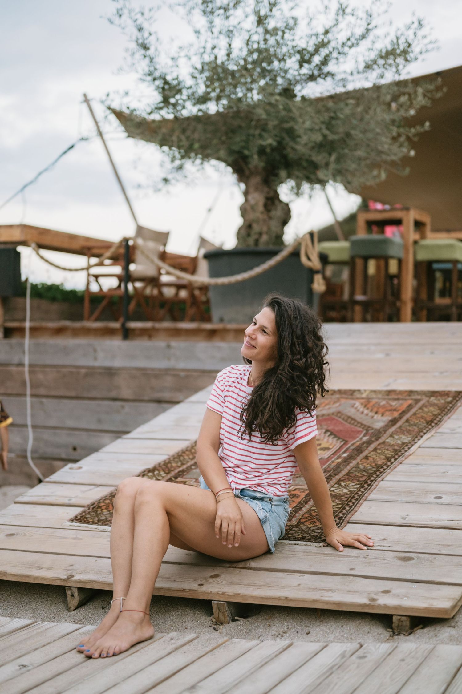 Young woman in striped tee and denim shorts sitting on wooden deck with patterned rug and olive tree backdrop.