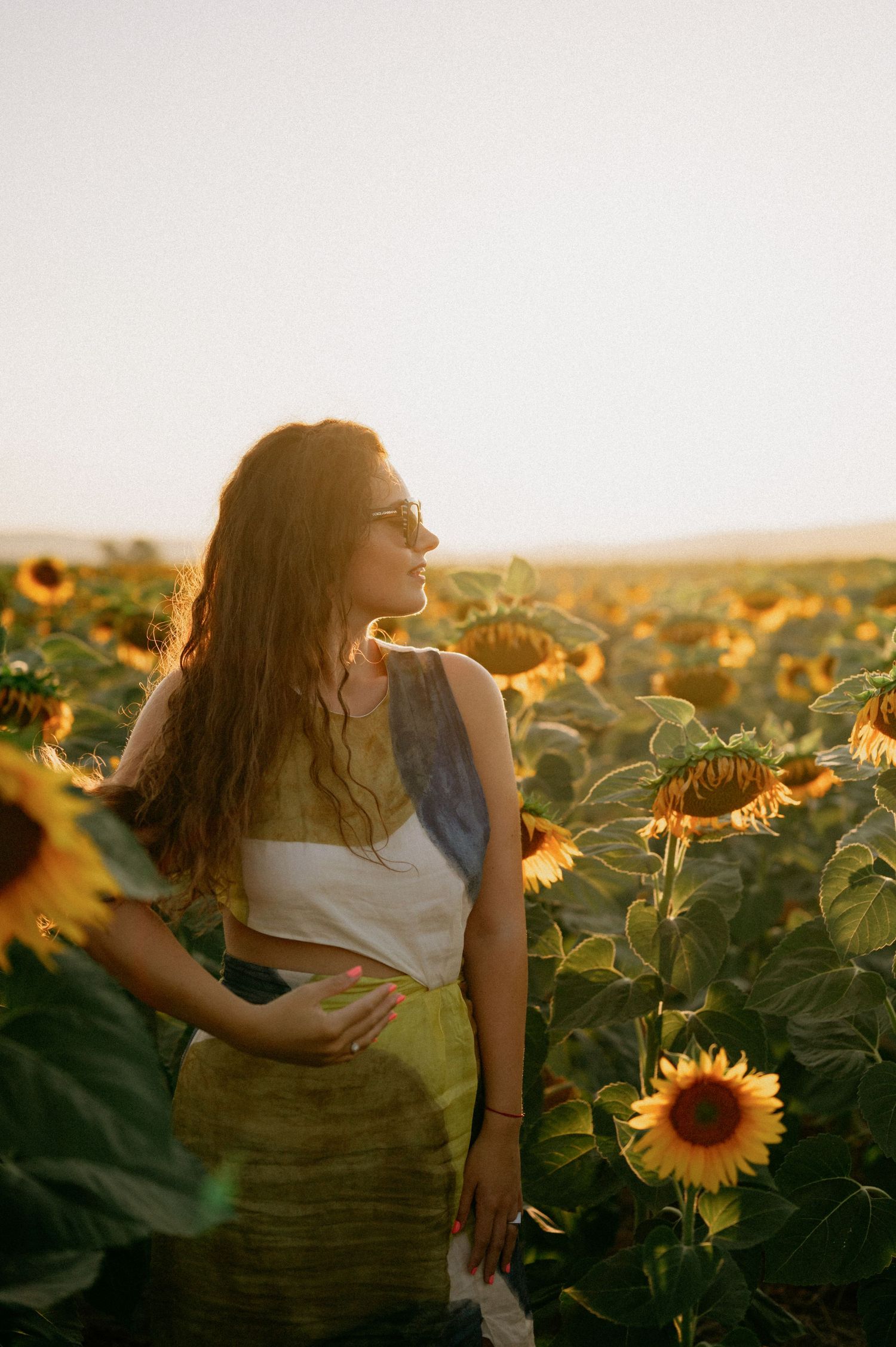 Woman standing in a sunflower field at golden hour, wearing a white top and green skirt, sunlight glowing behind her.