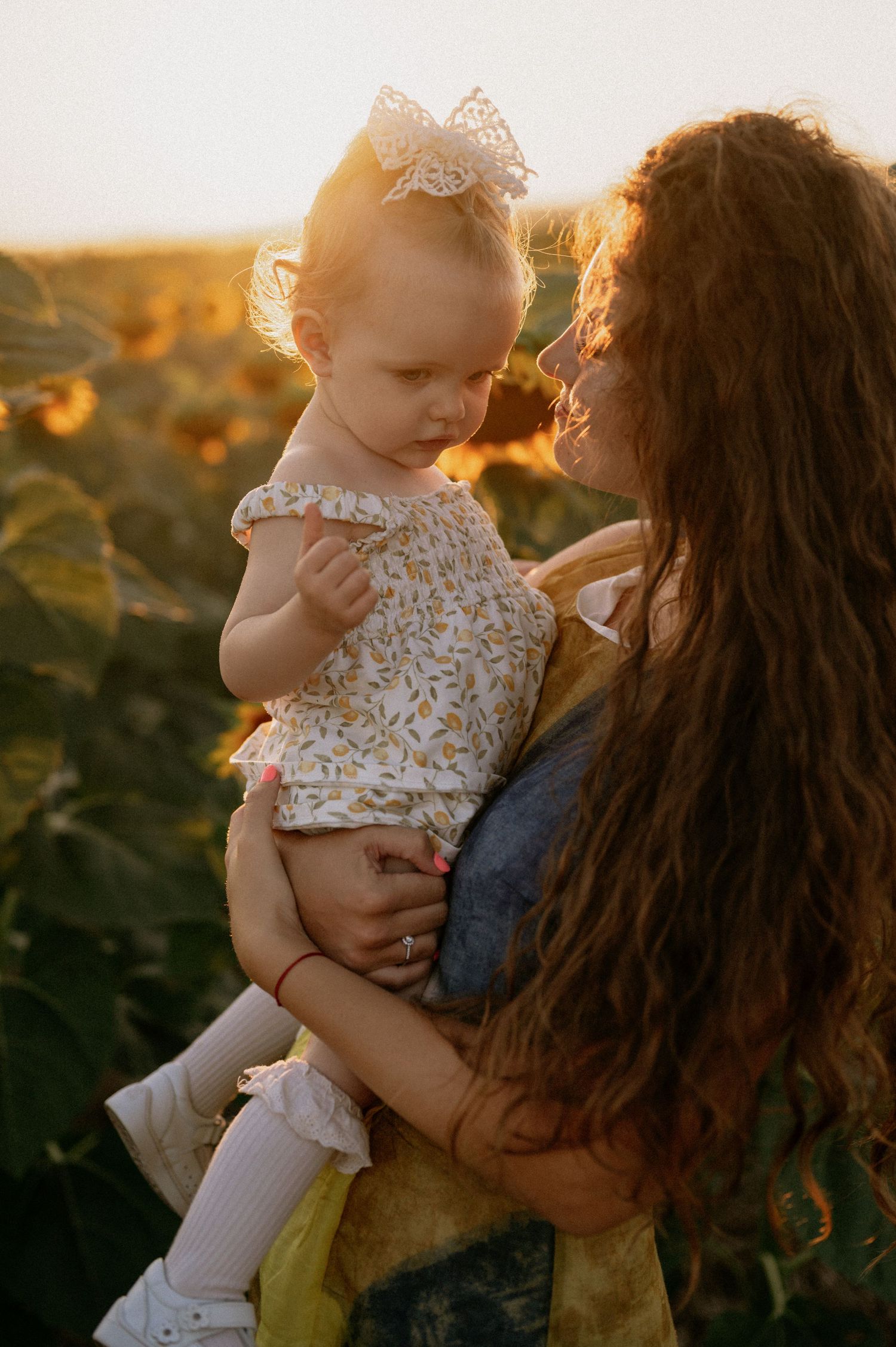 A mother with curly hair holds her baby girl in a sunflower field at golden hour sunset.
