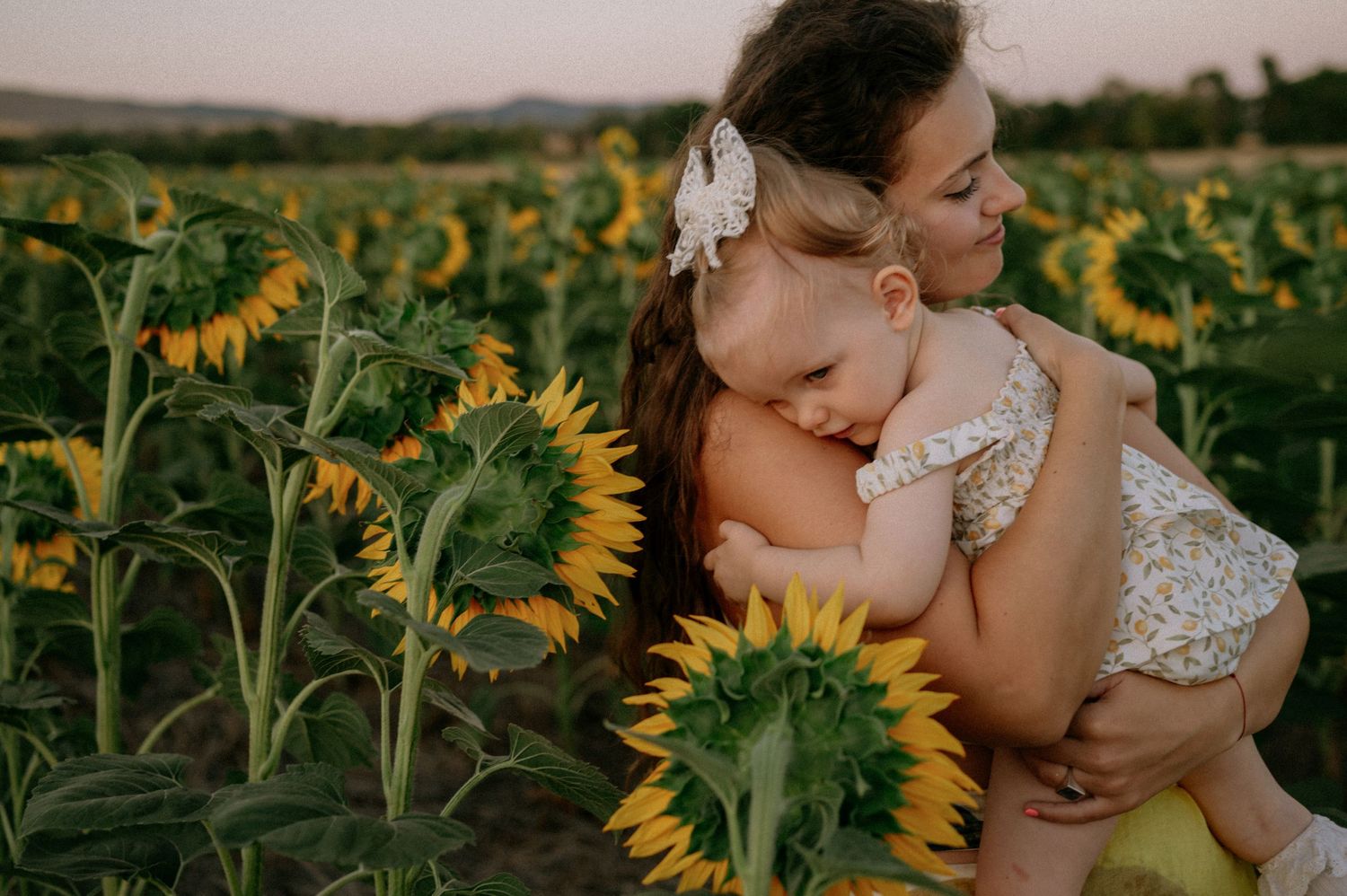 Mother holding sleeping baby in a sunflower field at golden hour, wearing floral outfit with white bow.