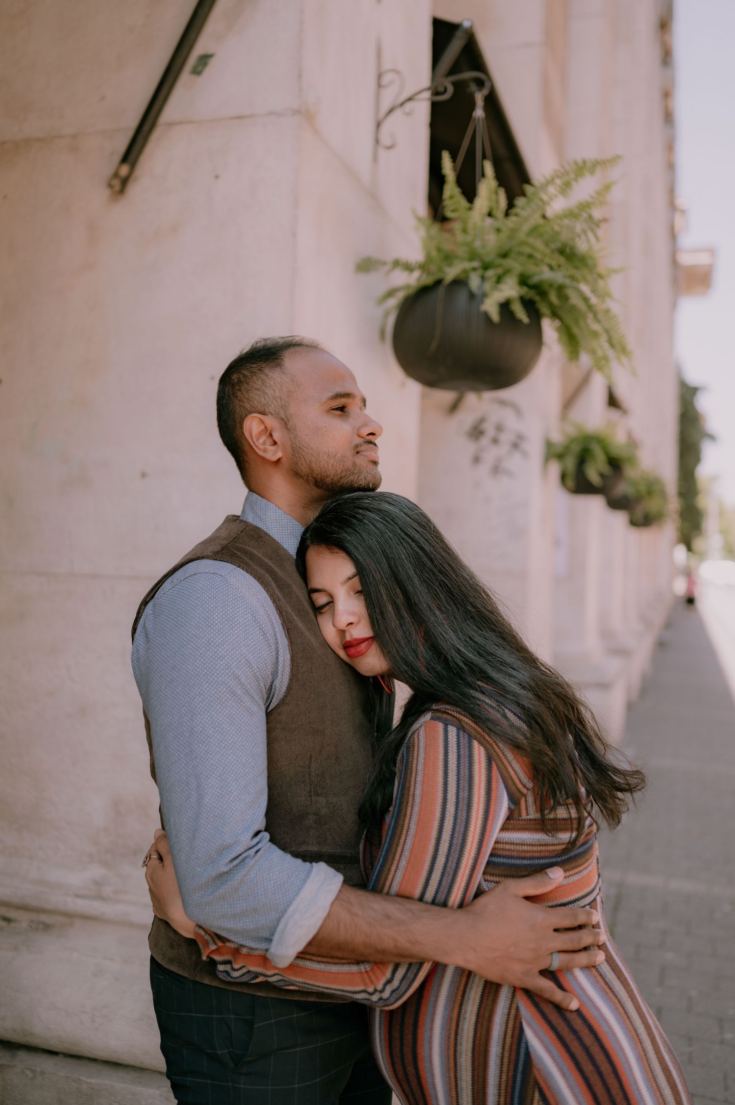 A couple embraces tenderly outside a building with hanging ferns, the woman resting her head on the man's chest.