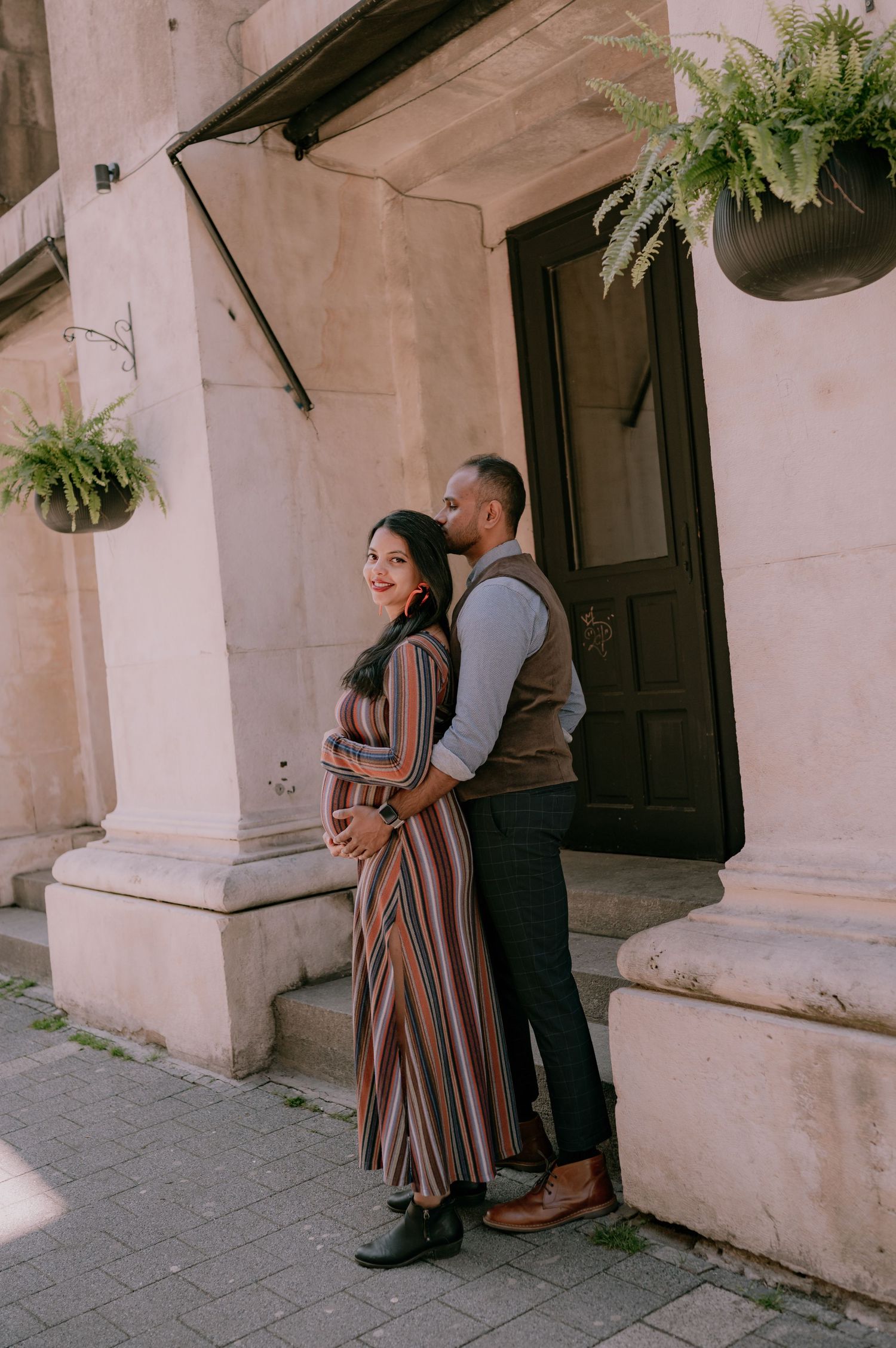 Pregnant couple embracing outside a historic building, woman in striped maxi dress smiling, man in vest kissing her head.