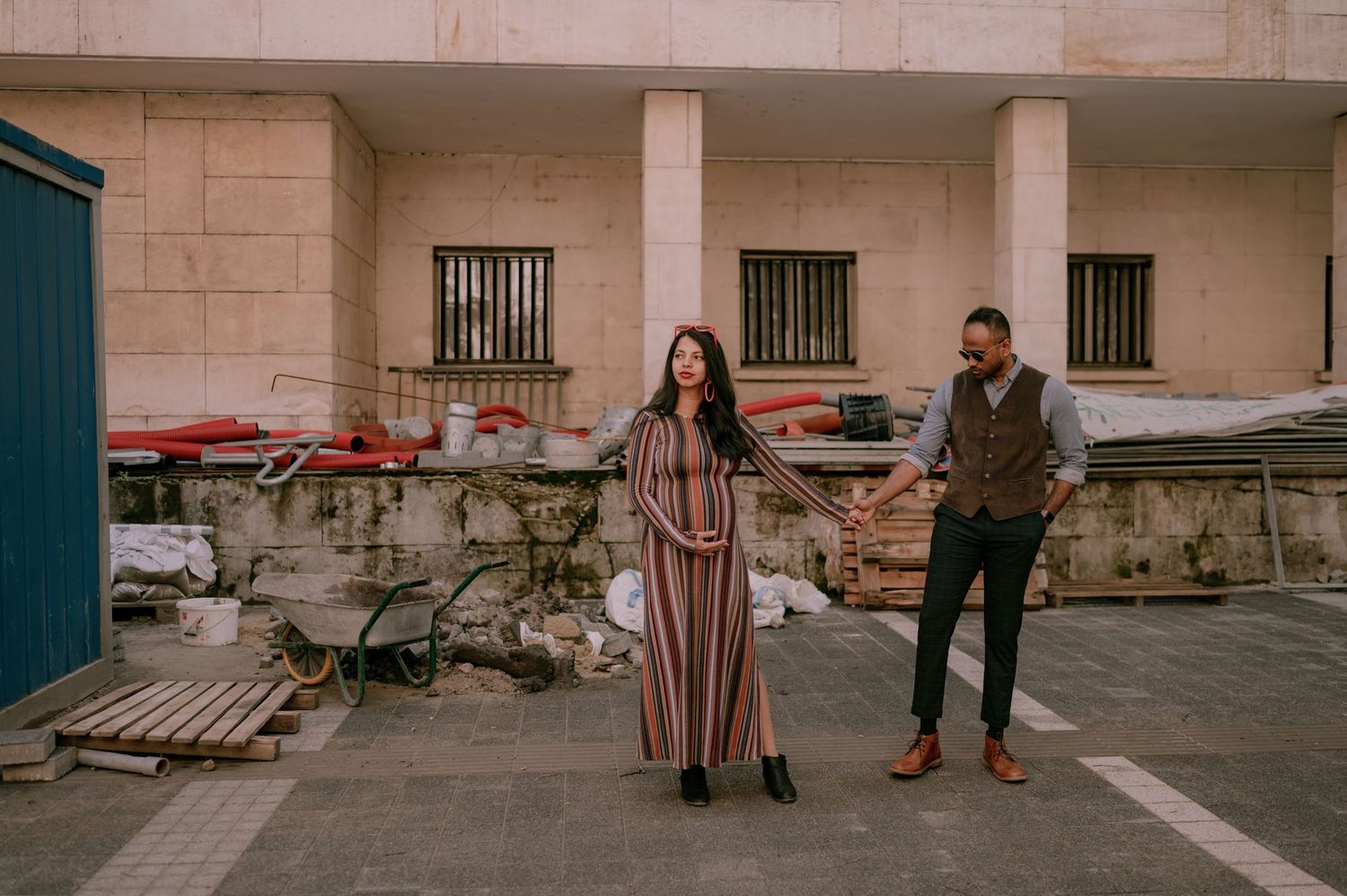 Couple holding hands in striped dress and dark suit posing in urban courtyard with rustic stone wall backdrop.
