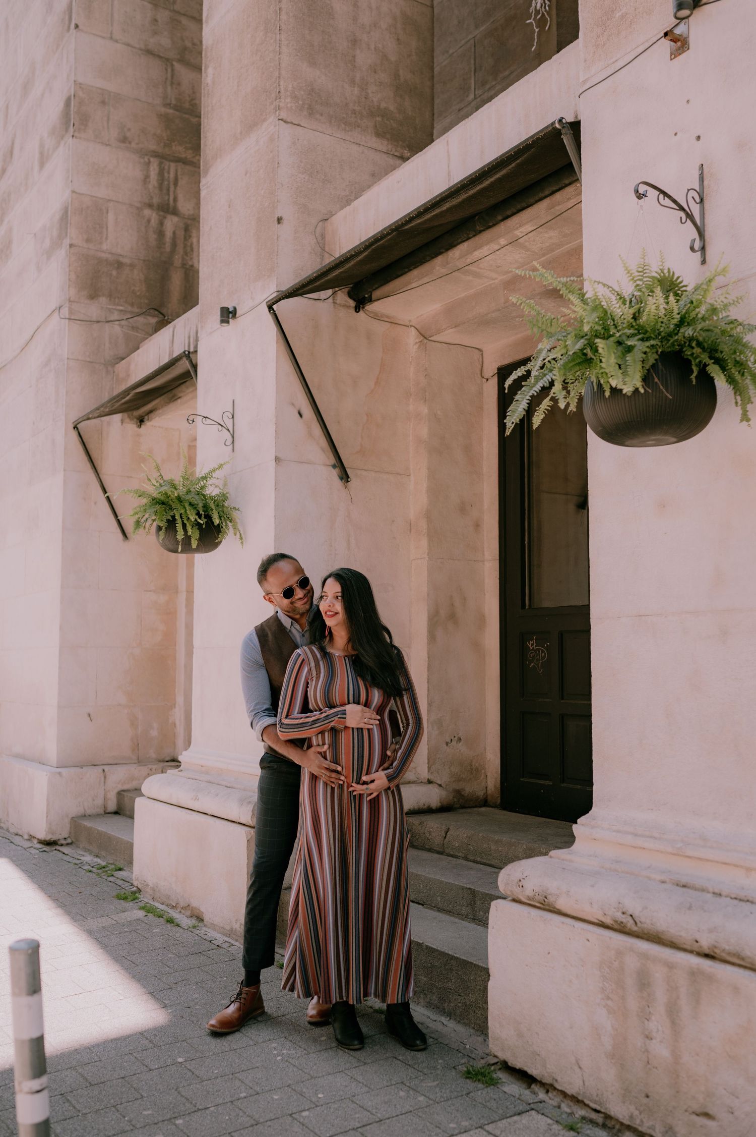 Couple sharing a tender moment outside a stone building with hanging ferns, woman in striped maxi dress cradling baby bump.