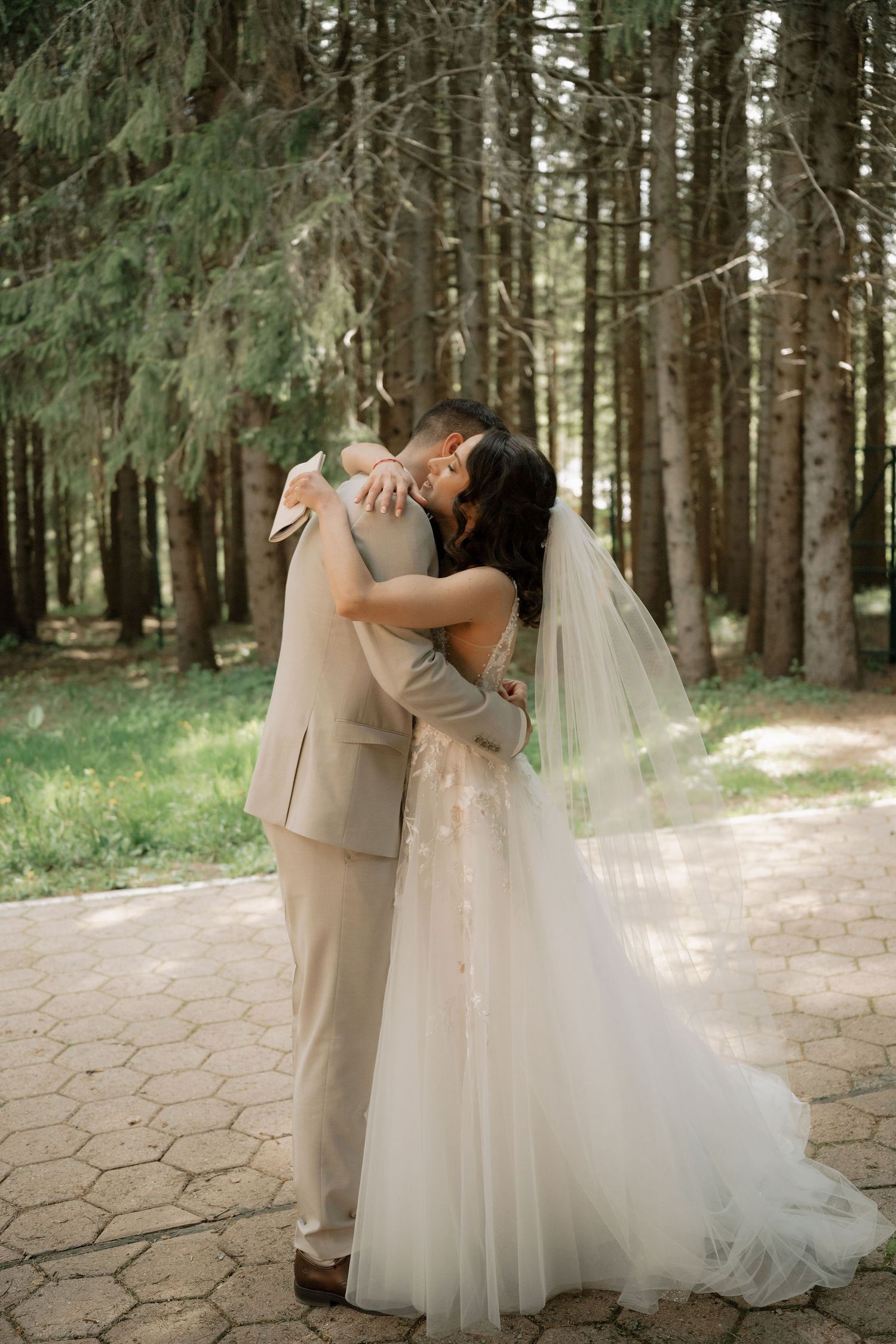 A bride and groom share a romantic embrace and kiss in a sunlit forest, with the bride wearing a flowing white tulle gown and veil.