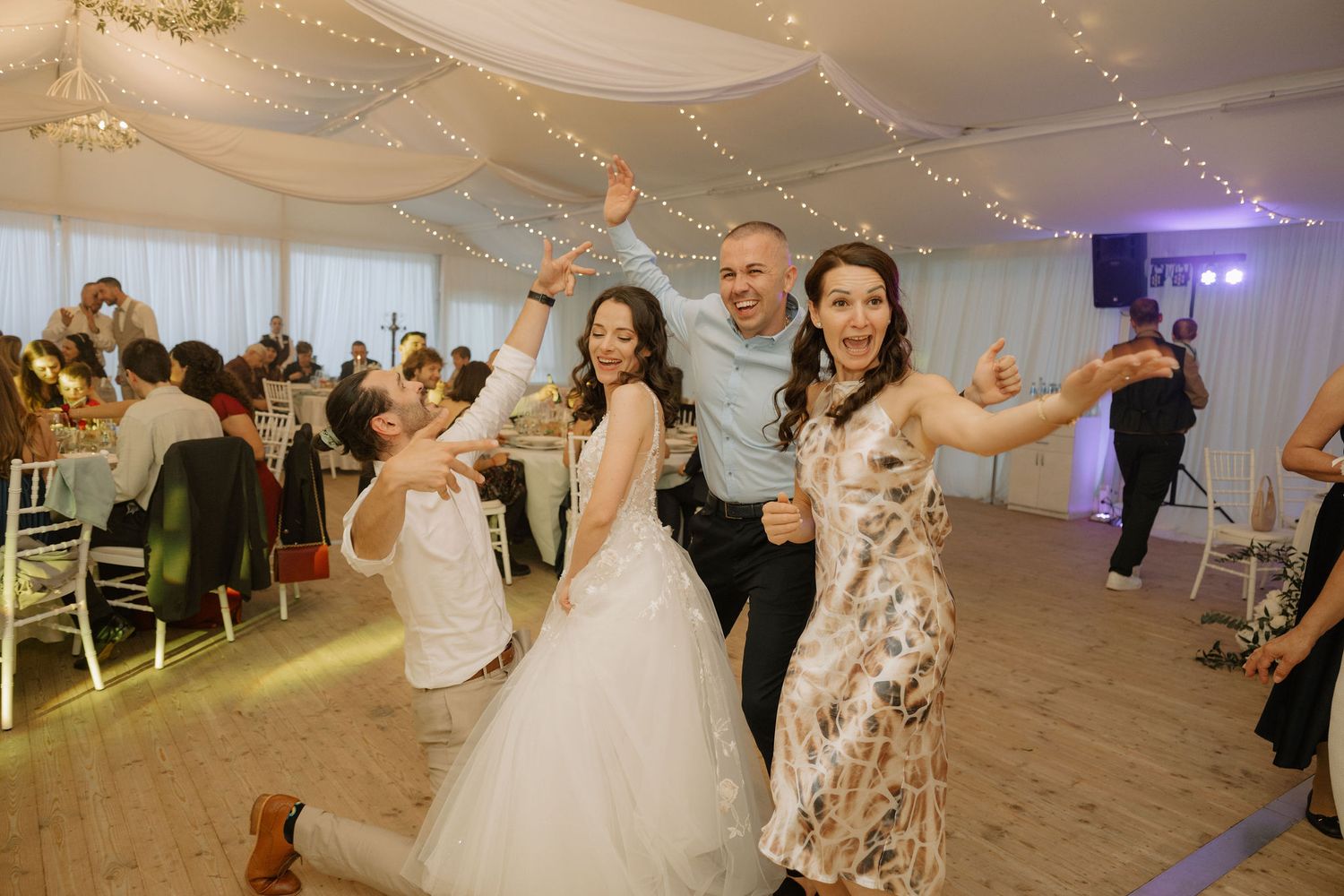 Bride in white gown celebrates with guests on wedding reception dance floor under fairy lights.