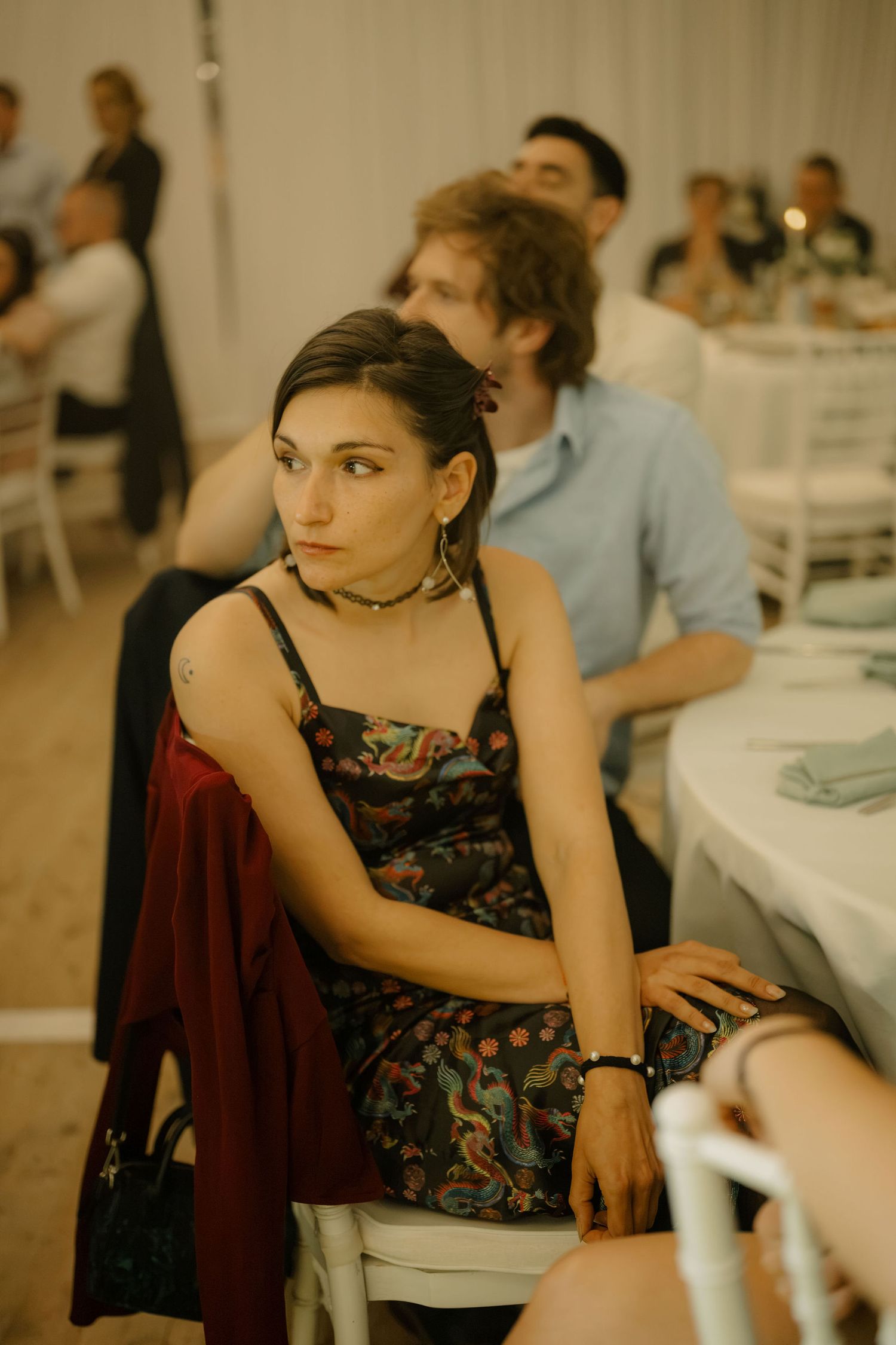 Woman in floral dress sitting at wedding reception table, wearing choker necklace and gold earrings, looking away thoughtfully.