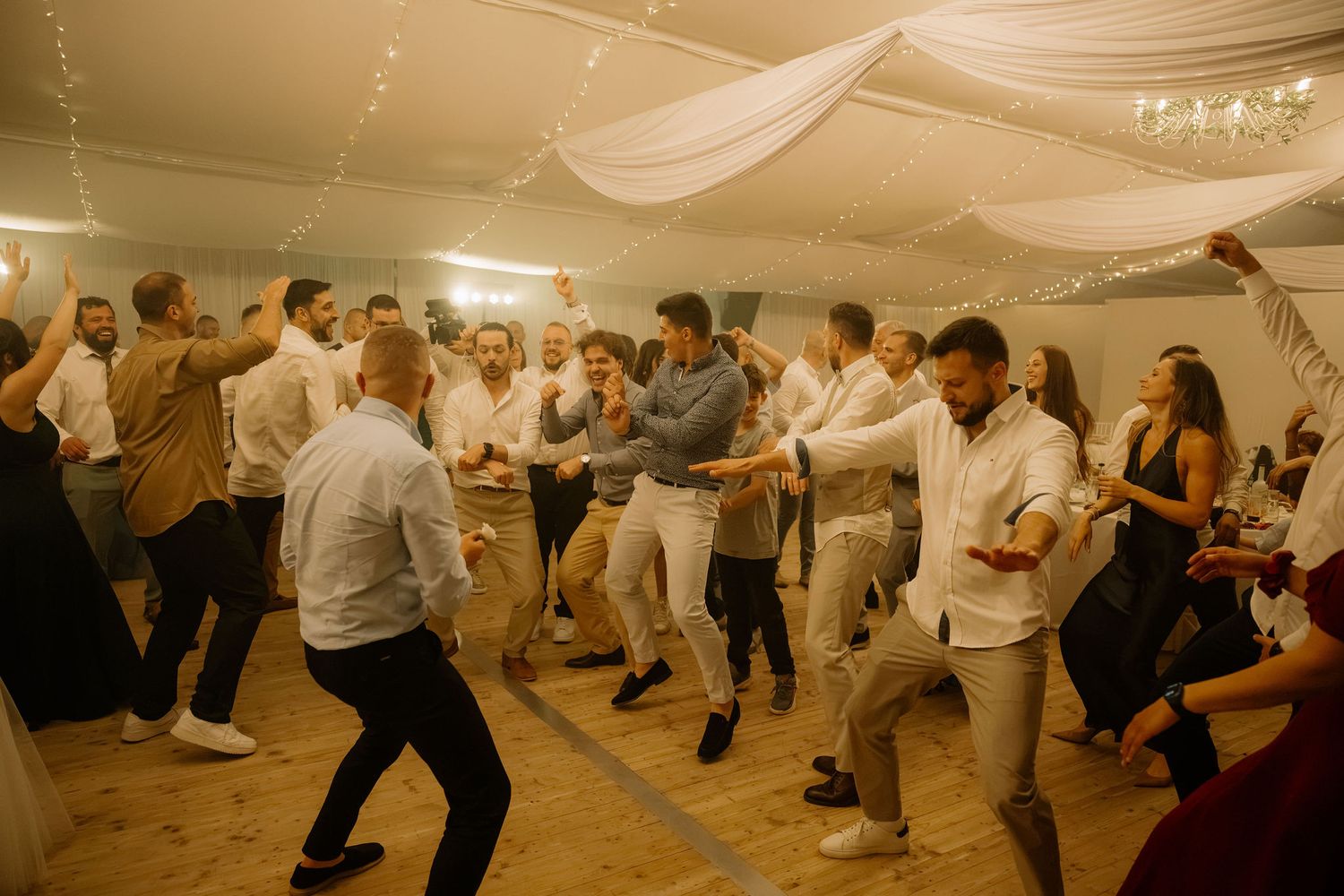 Guests dancing energetically at a wedding reception in a decorated marquee tent with draped white fabric ceiling.