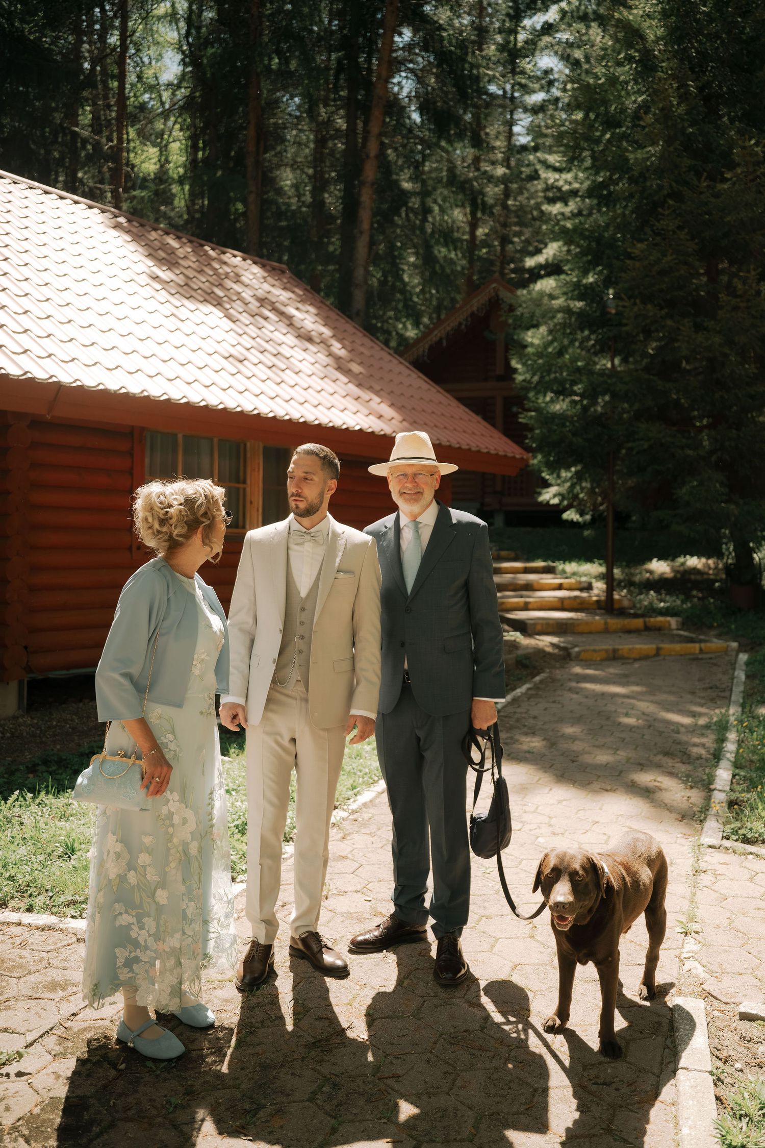 Groom in beige suit poses with elderly parents and brown Labrador dog outside rustic wooden cabin in forest setting.