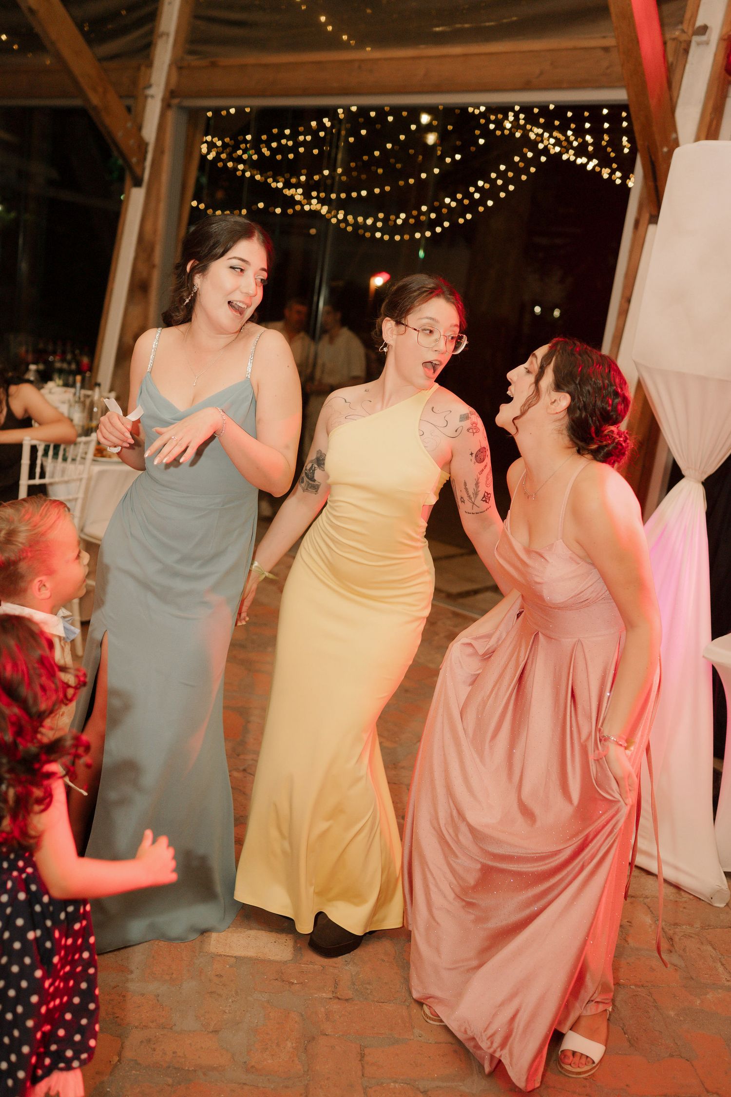 Three women in elegant formal dresses dance and laugh together at an event decorated with string lights.