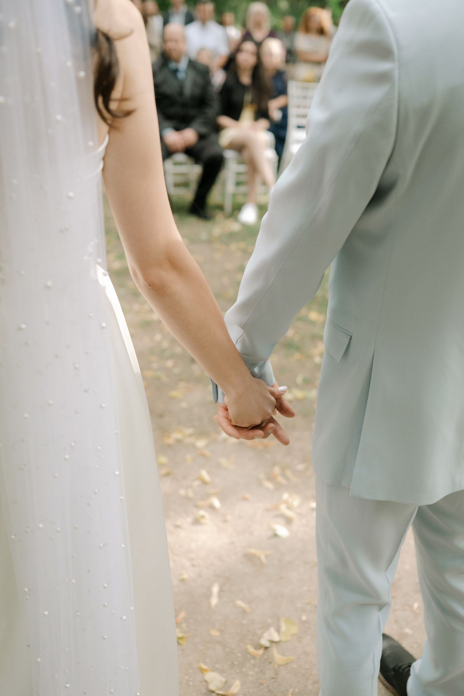 A couple in wedding attire holding hands outdoors with guests visible in the background during a ceremony.