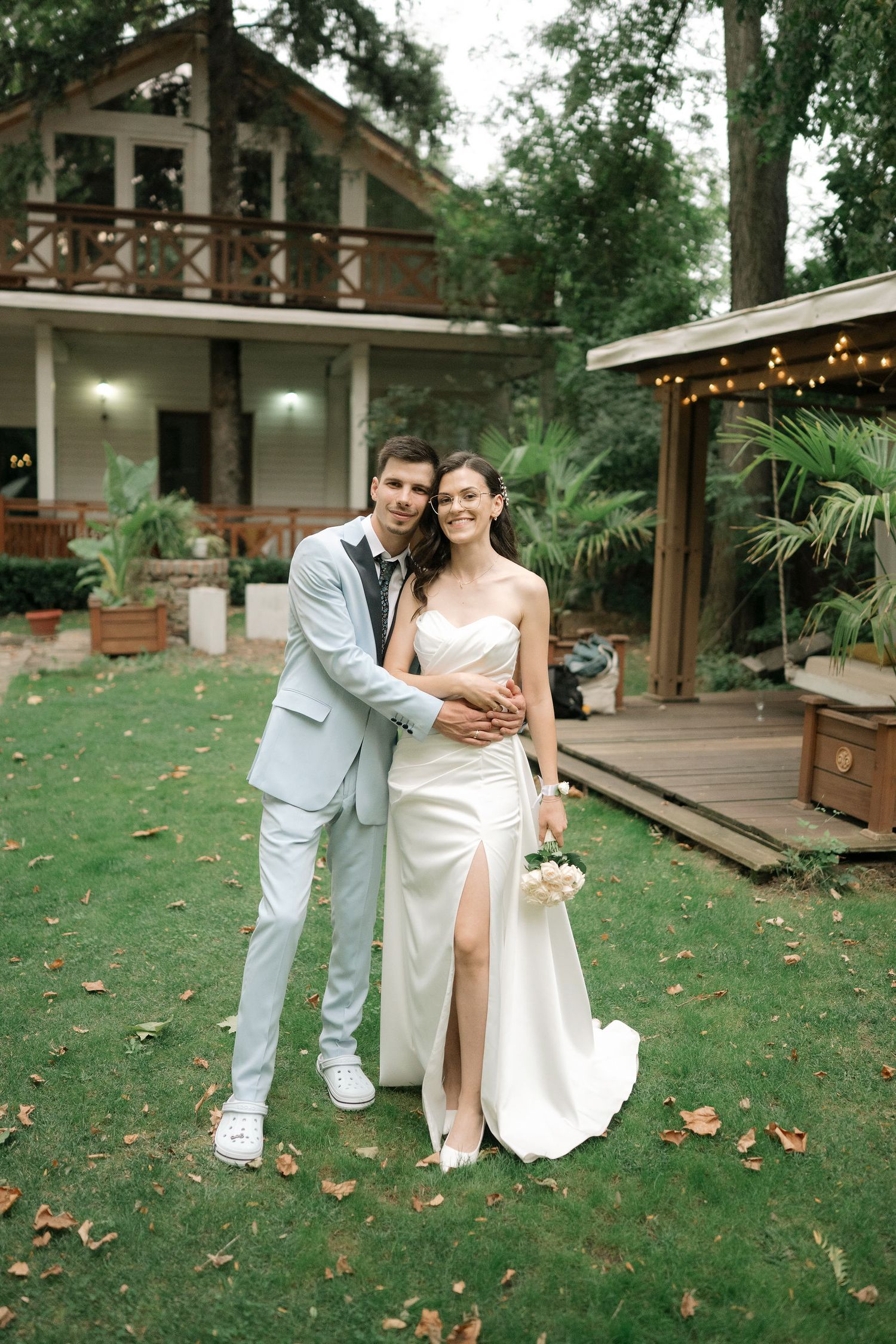 A couple poses together on a grassy lawn in front of a rustic wooden building with string lights at dusk.