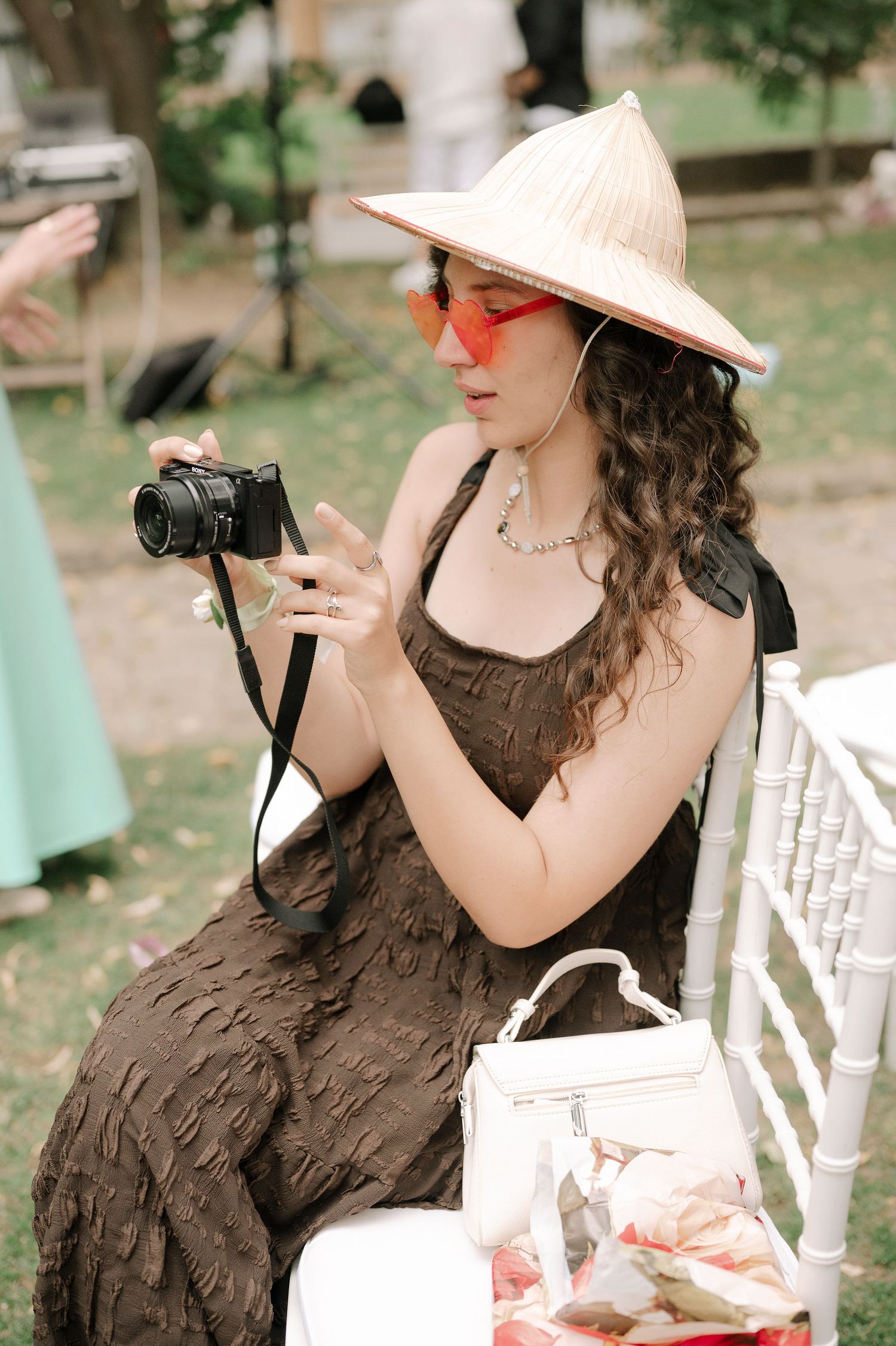 Woman in brown dress and sun hat poses elegantly in outdoor garden setting with white chairs.