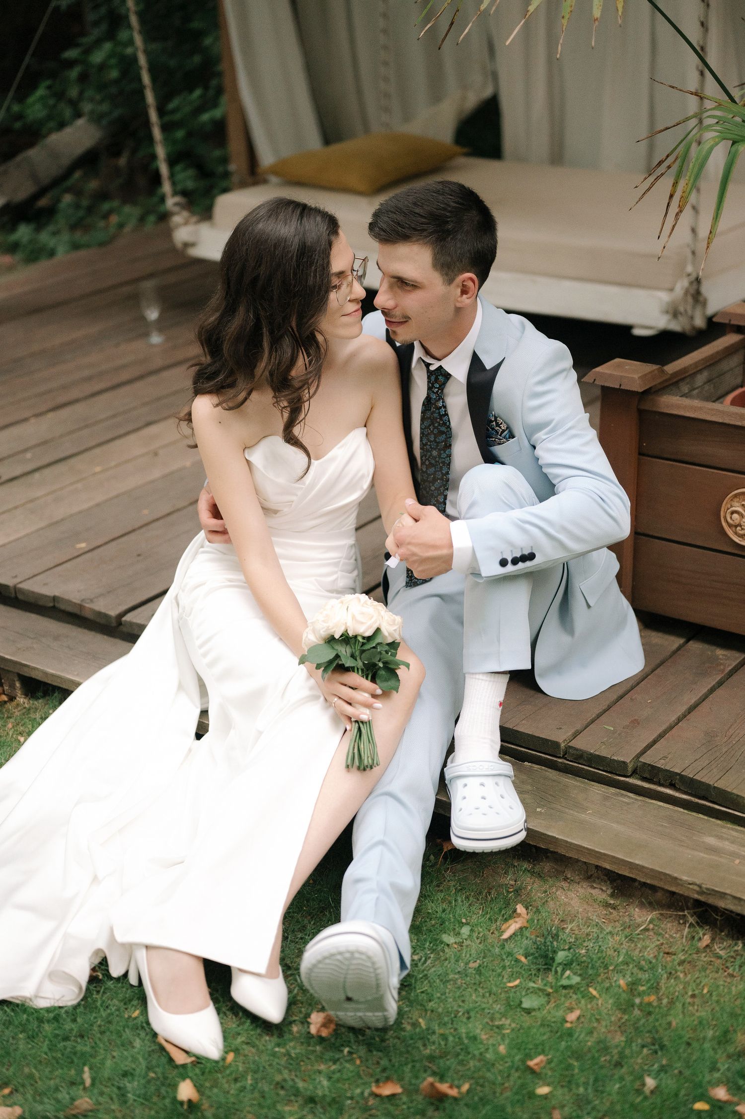 A romantic wedding couple sits together on wooden deck steps surrounded by greenery.