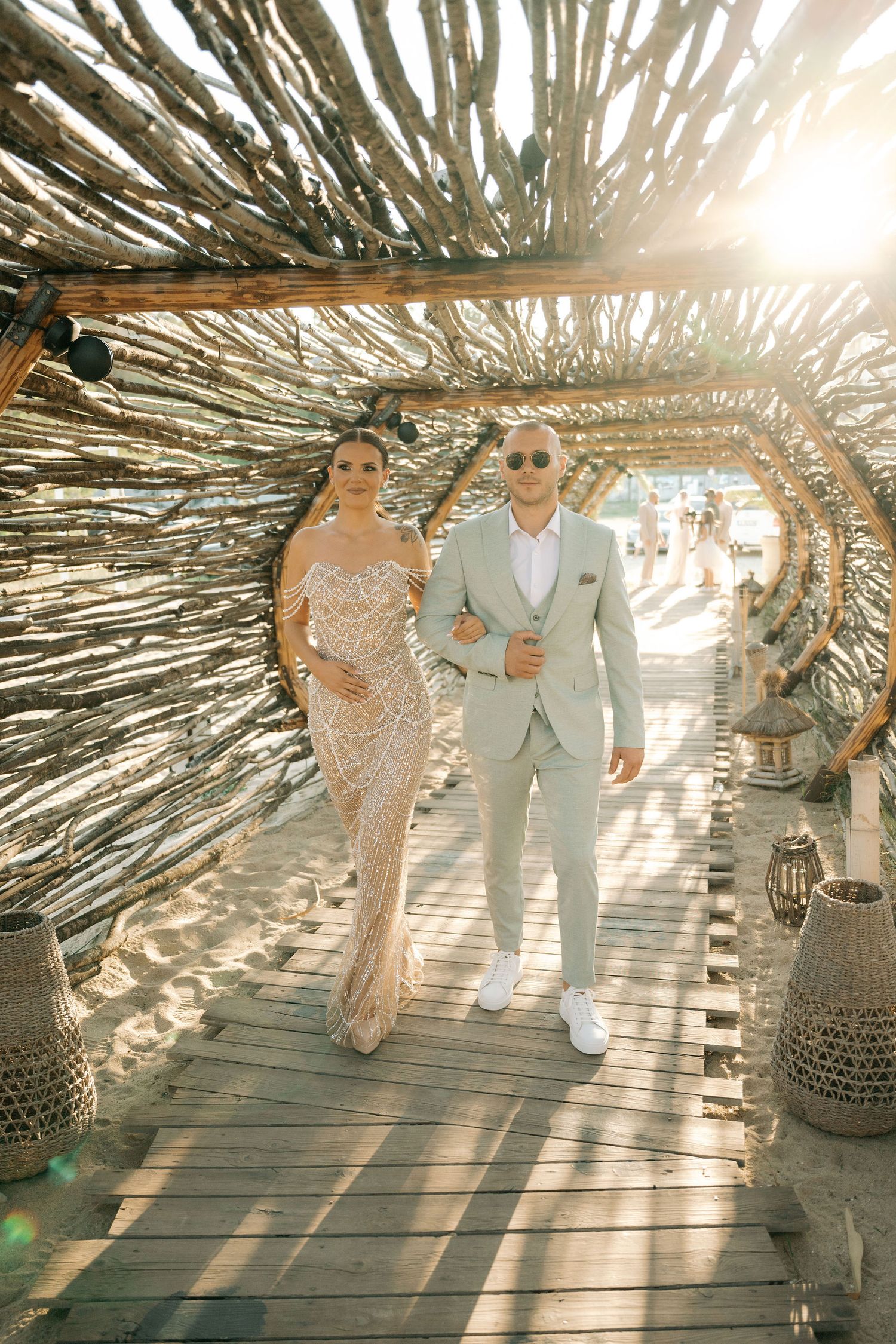People walk together on a wooden deck under a natural palm frond canopy creating dramatic sunlit patterns.