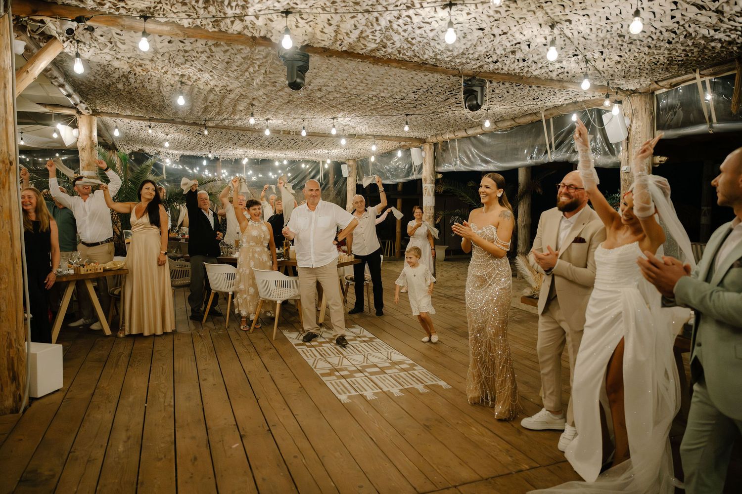 Wedding reception guests dancing and celebrating under string lights on a rustic wooden deck at an evening party.