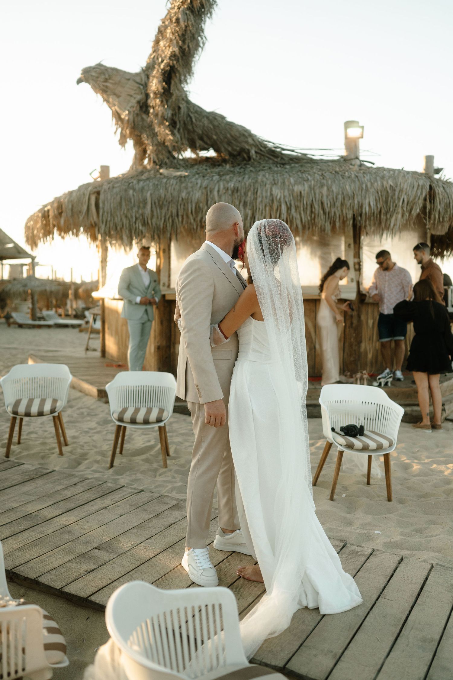 Beach wedding reception under thatched palapa with white chairs and wooden deck at golden hour.