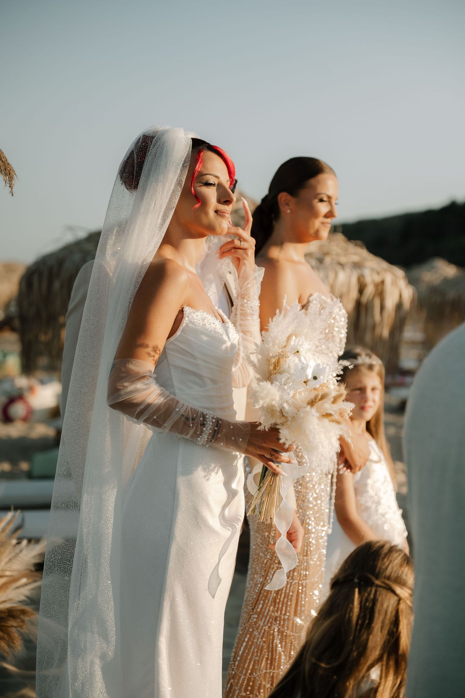 Two women in elegant white dresses pose together against a scenic coastal backdrop during golden hour.