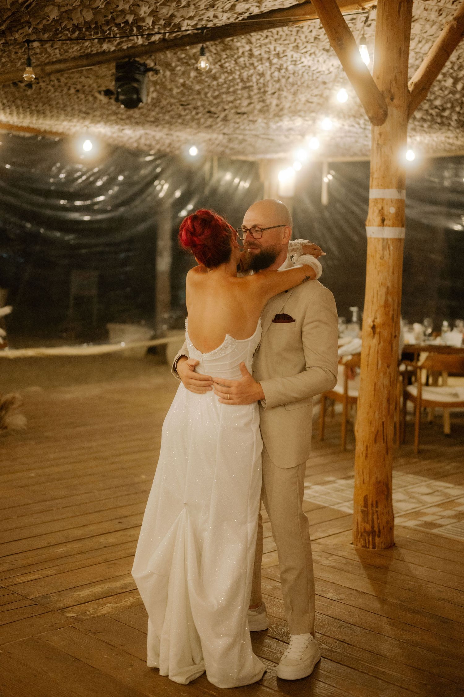A couple shares a romantic slow dance on a rustic wooden dance floor under warm glowing lights at their wedding reception.
