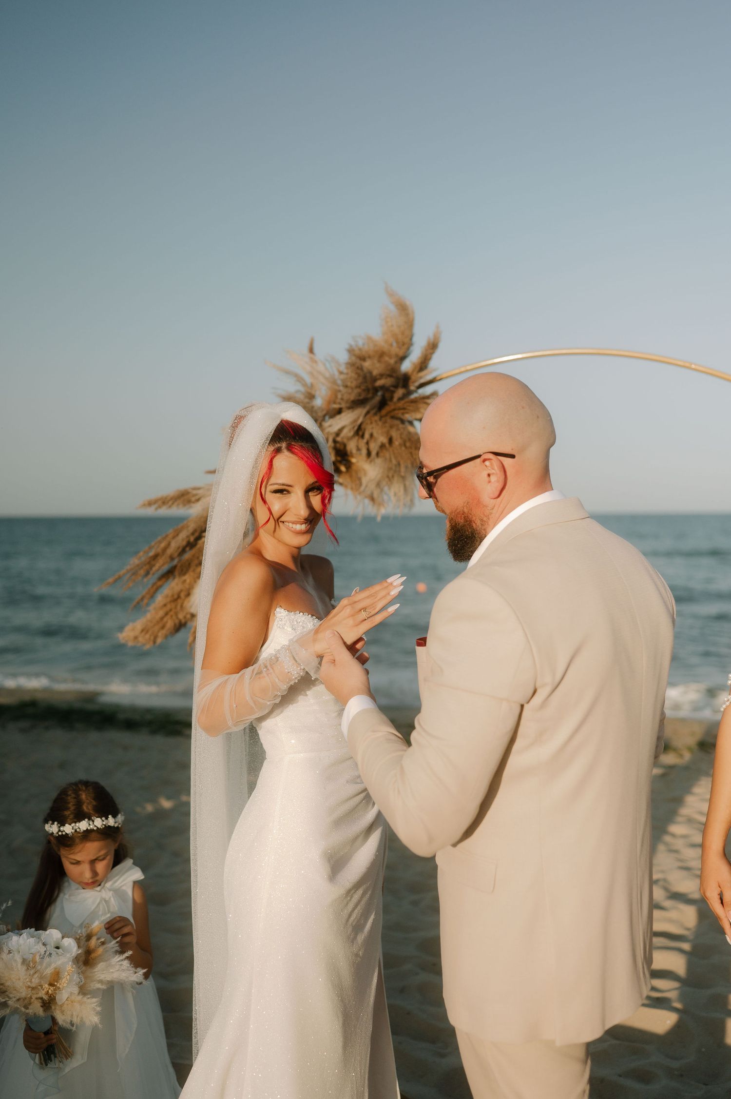 A couple shares a joyful moment dancing together on a beach at sunset in matching light-colored outfits.